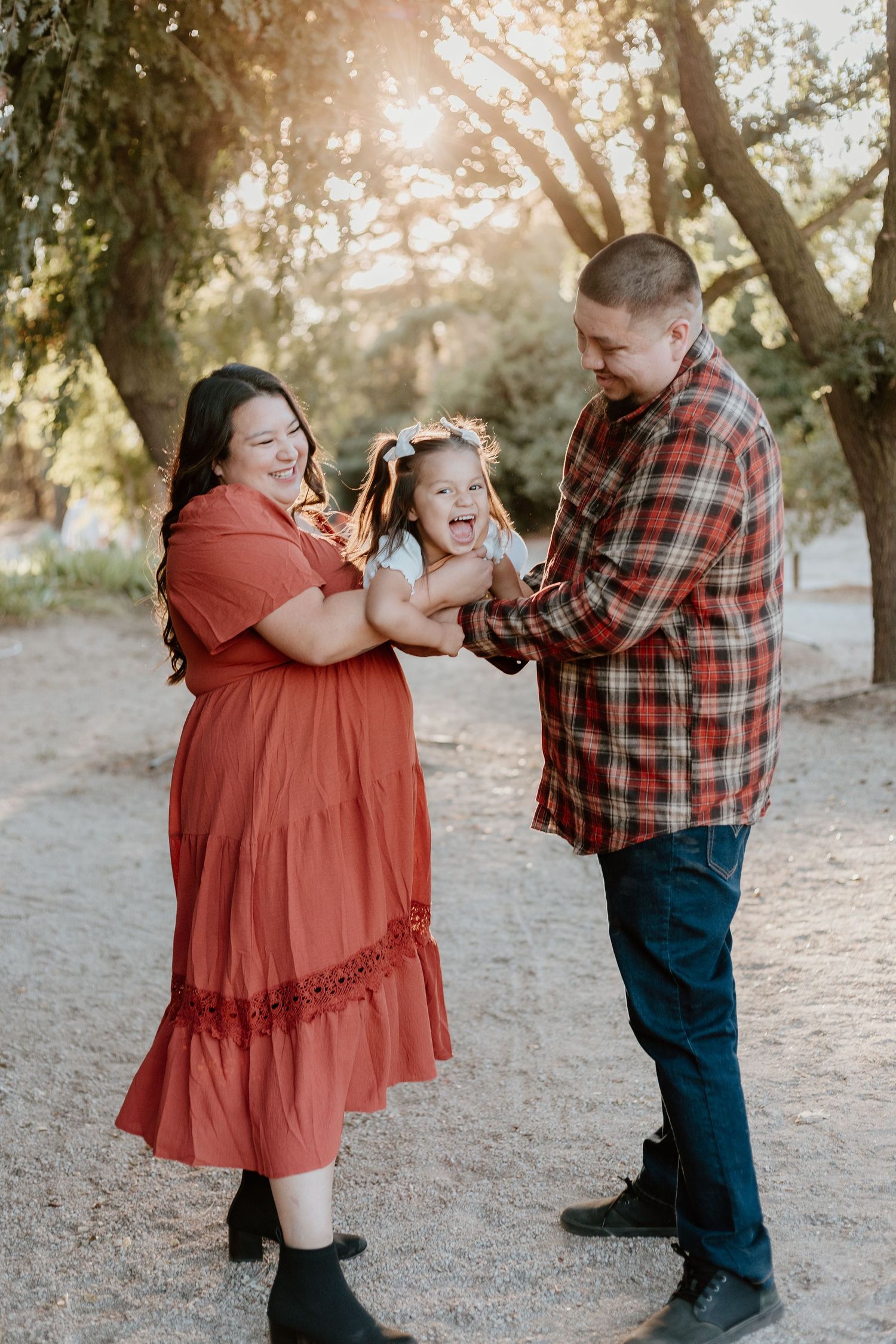 A joyful family of three plays outdoors at golden hour, mom in red dress and dad in plaid holding their laughing toddler.