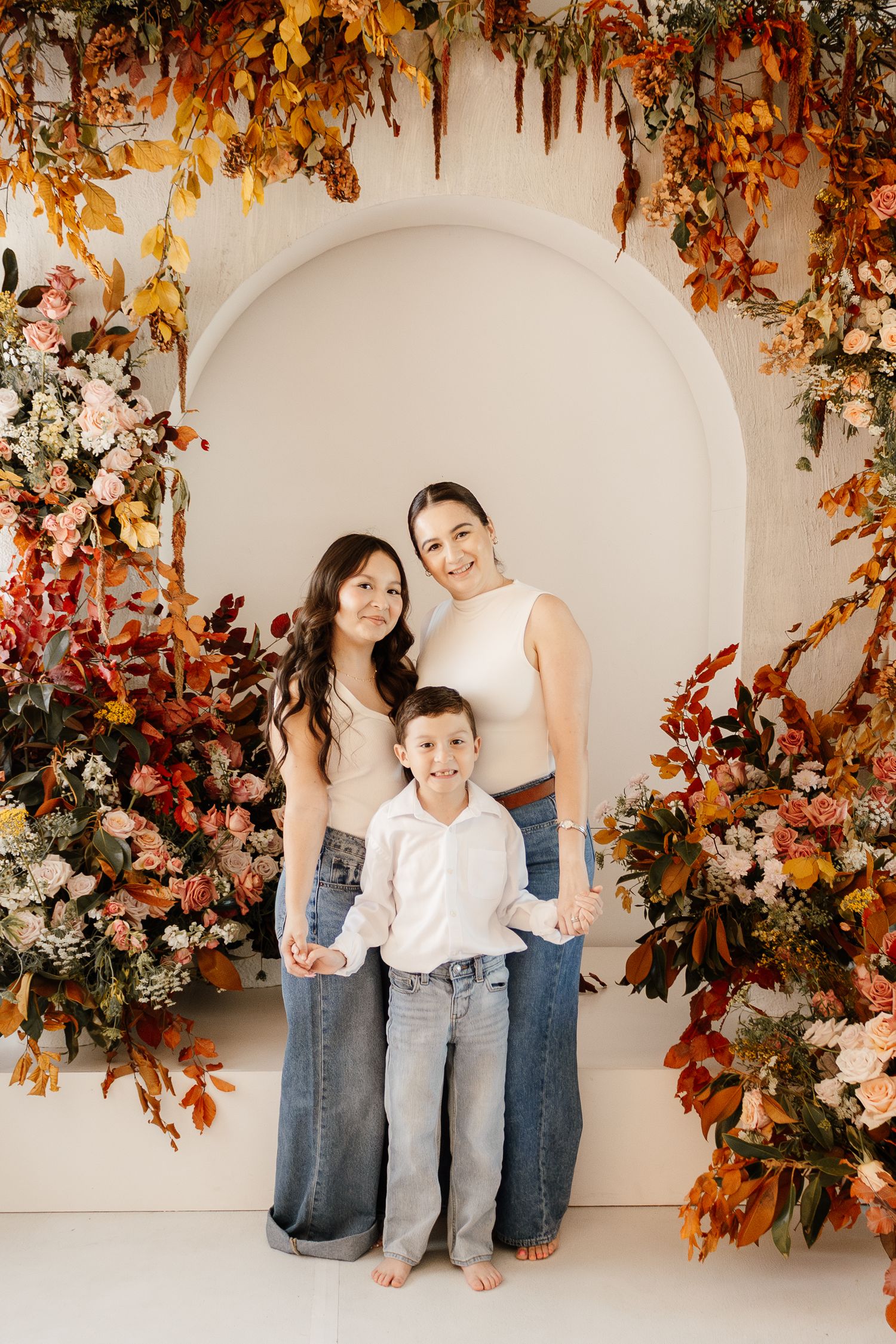 Family of three posing in front of floral arch with autumn leaves and roses in a bright studio setting.