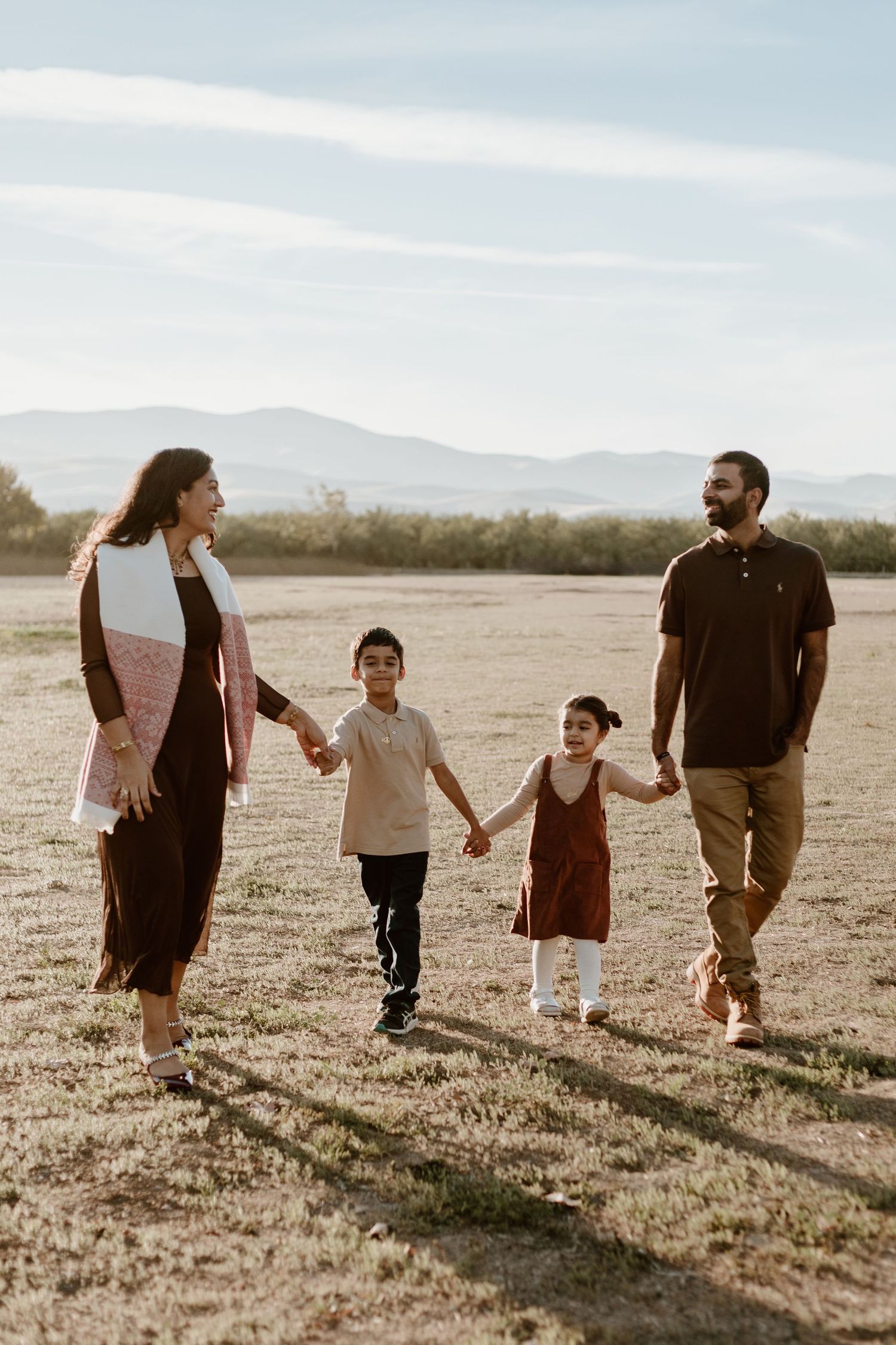 A family of four holds hands while walking through an open field with mountains in the background during golden hour.