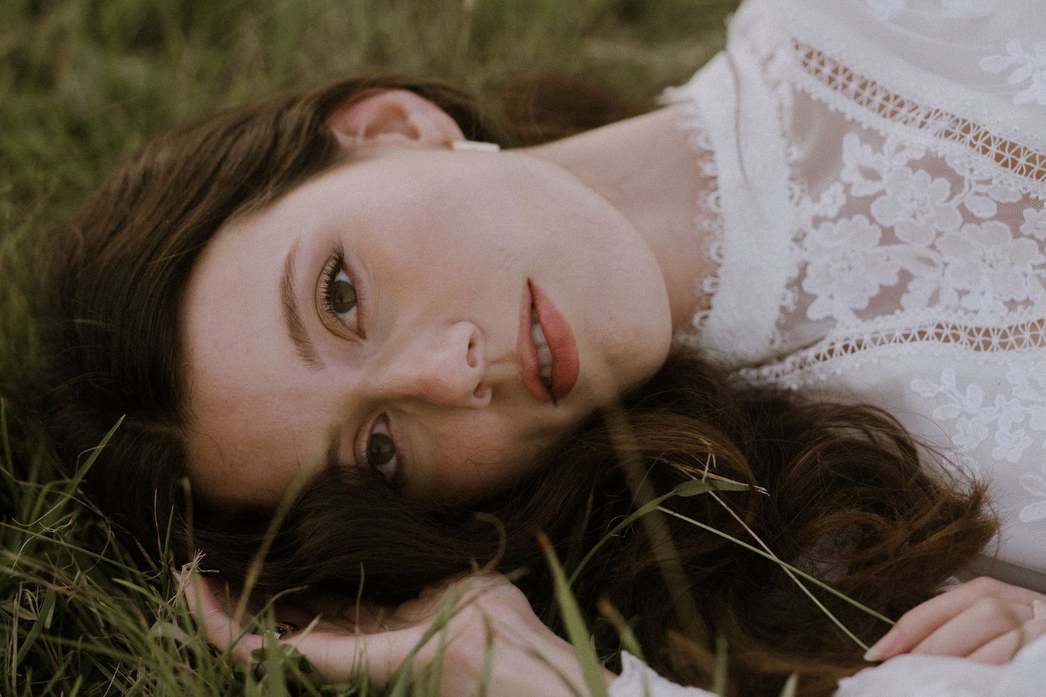A dreamy portrait of someone lying in grass wearing a white lace top with long dark hair.