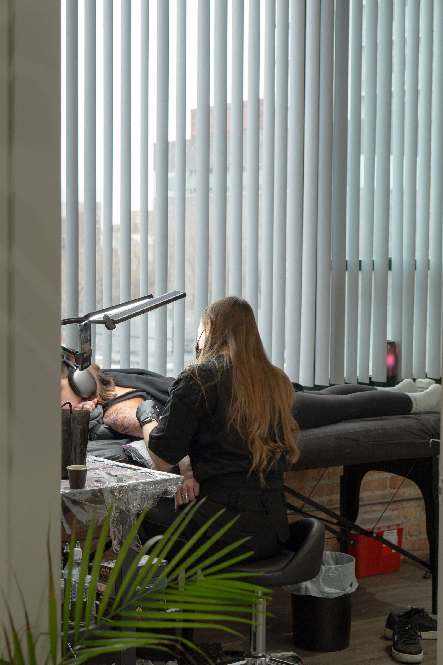 Tattoo artist works on a client in a professional studio setting with natural light filtering through vertical blinds.
