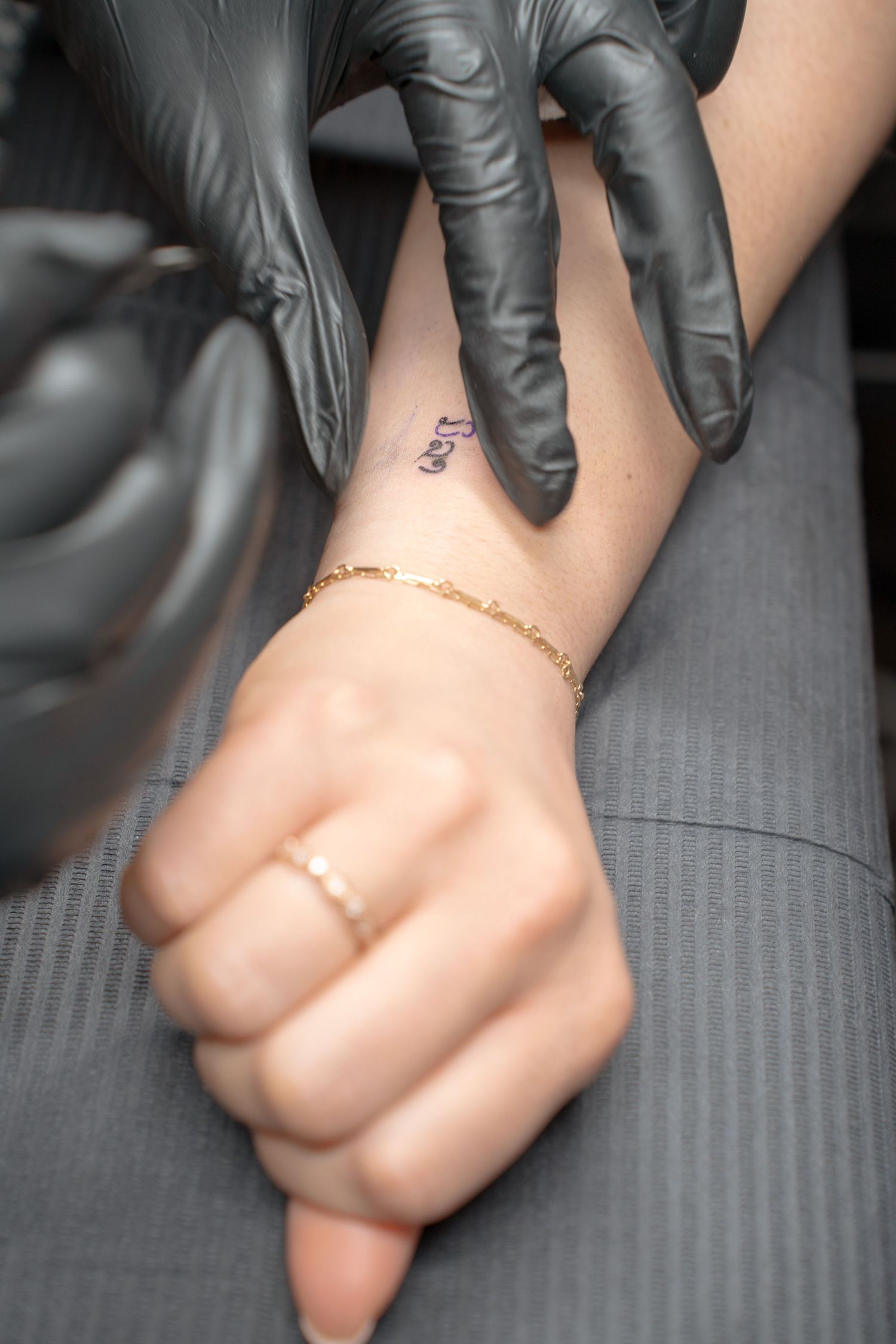 Close-up of gloved hands working on small tattoo with delicate gold bracelet visible.