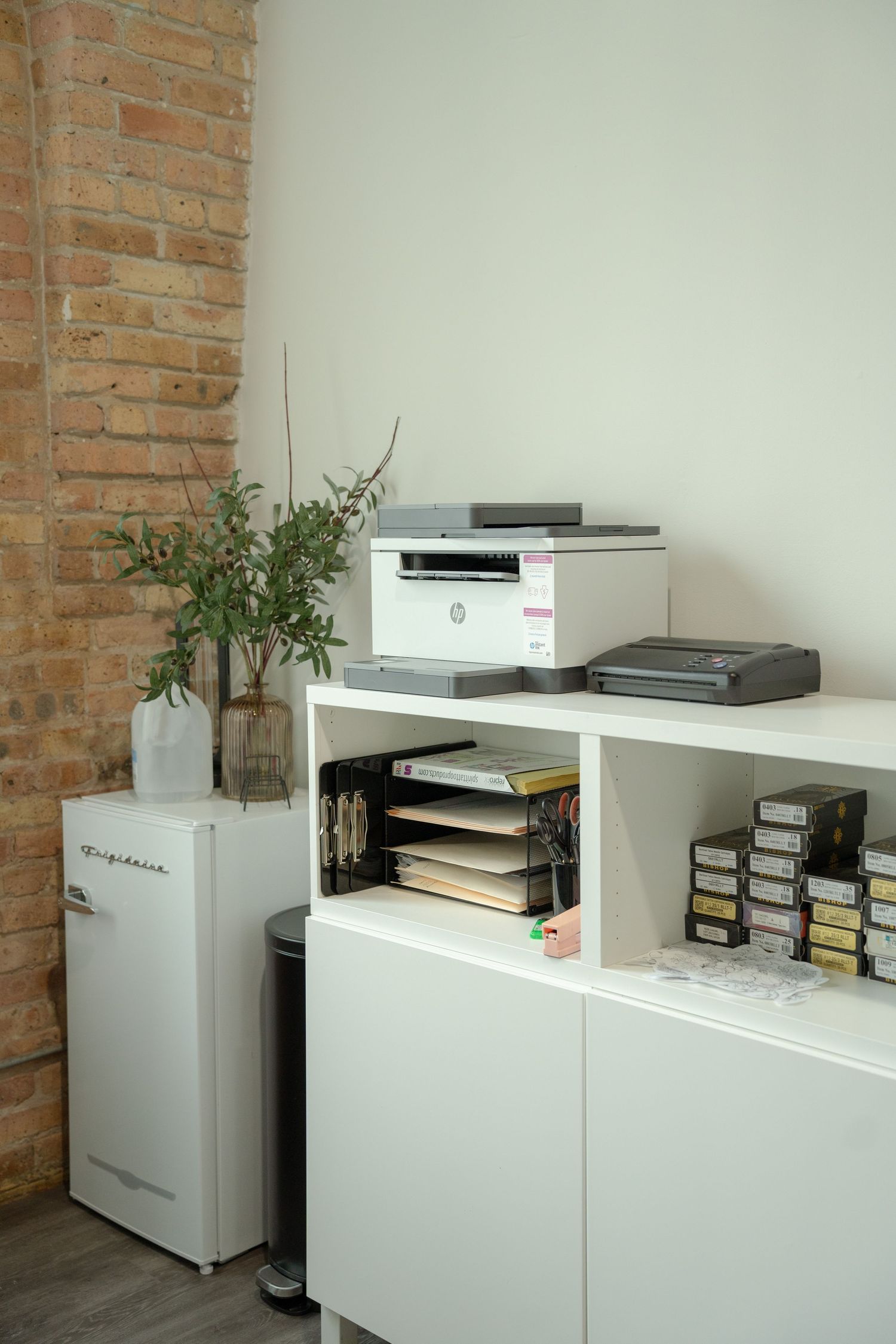 White office storage cabinet with printer and indoor plant against exposed brick wall.