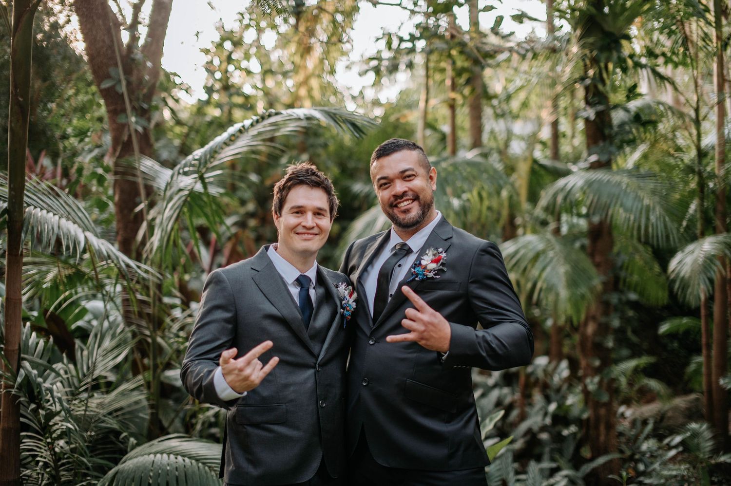 Two groomsmen in dark suits pose playfully together in a tropical garden setting.