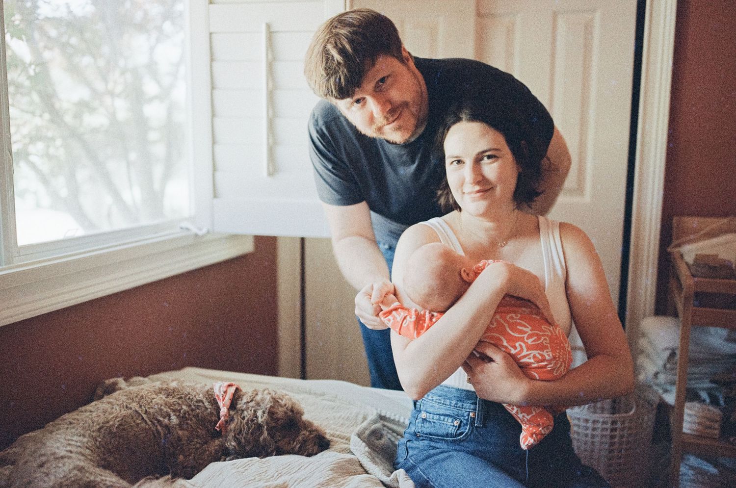 A family sits together on a bed with their cat and baby in a sunlit room.