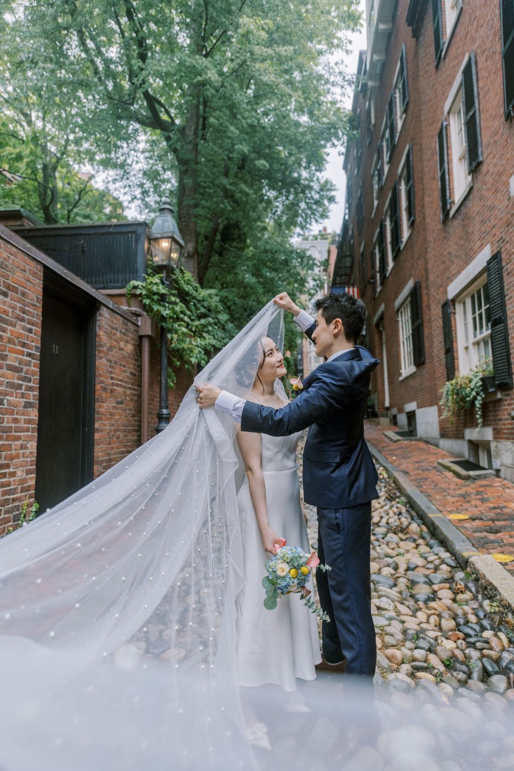 Couple posing with flowing veil on cobblestone street with brick buildings.