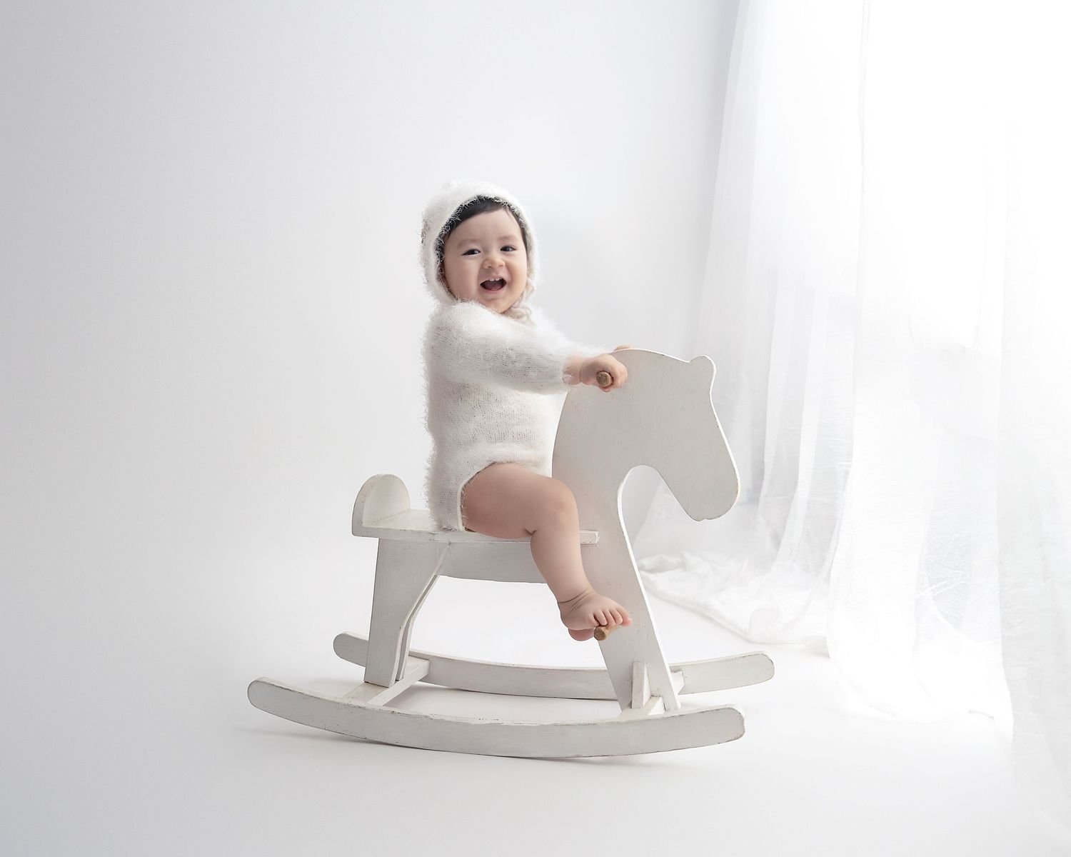 Baby plays on white rocking horse against bright minimalist studio background.
