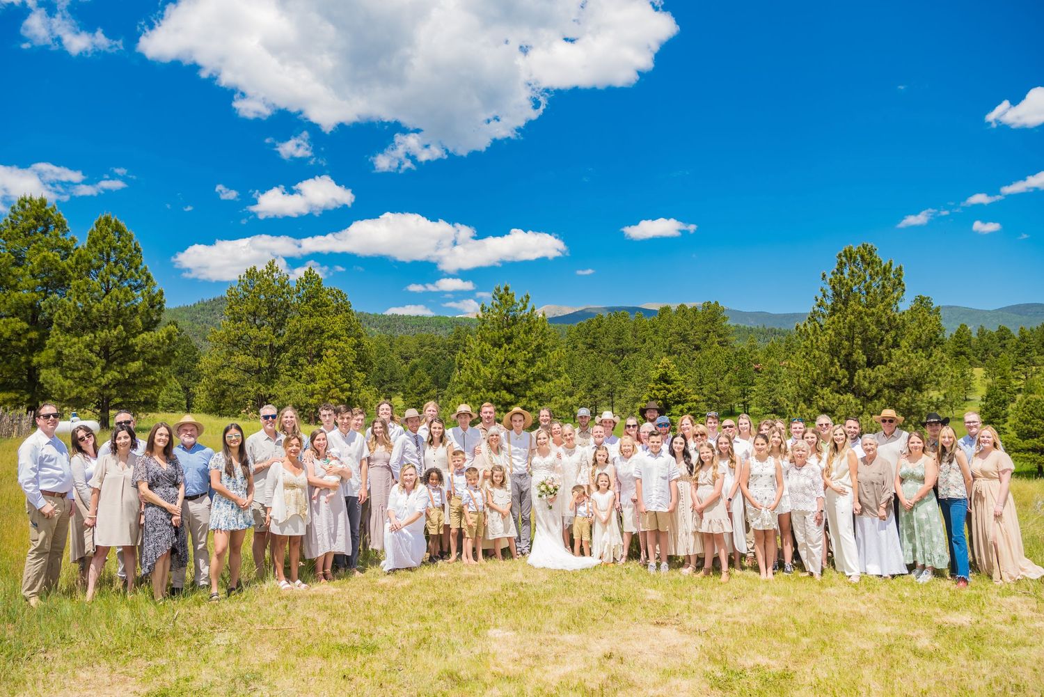 Large wedding group photo of guests wearing white and light colored attire in a scenic mountain setting.