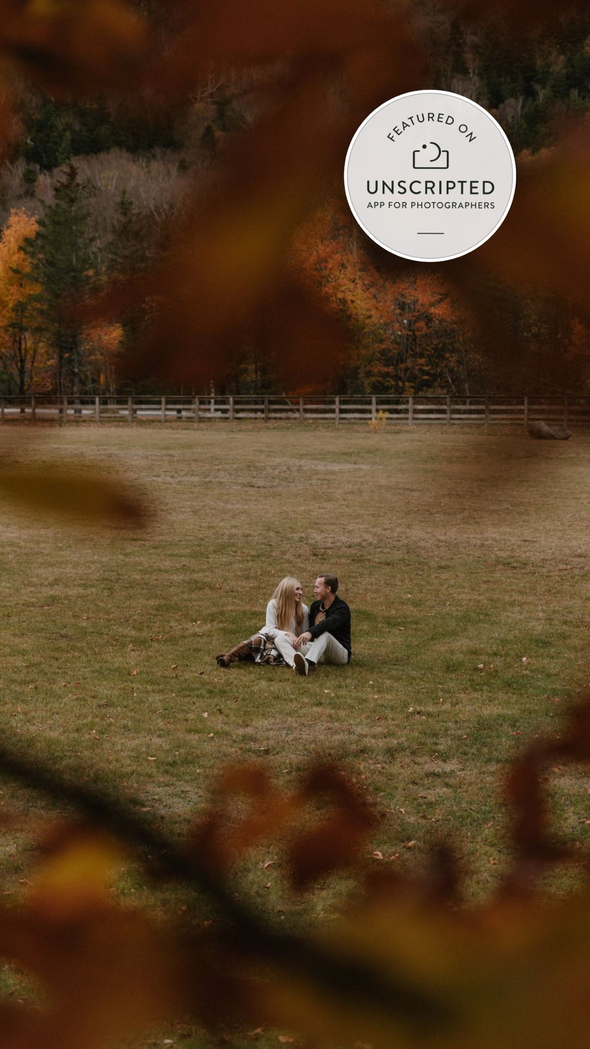 A couple sits together on a blanket in an autumn field with warm orange and brown tones in the background.