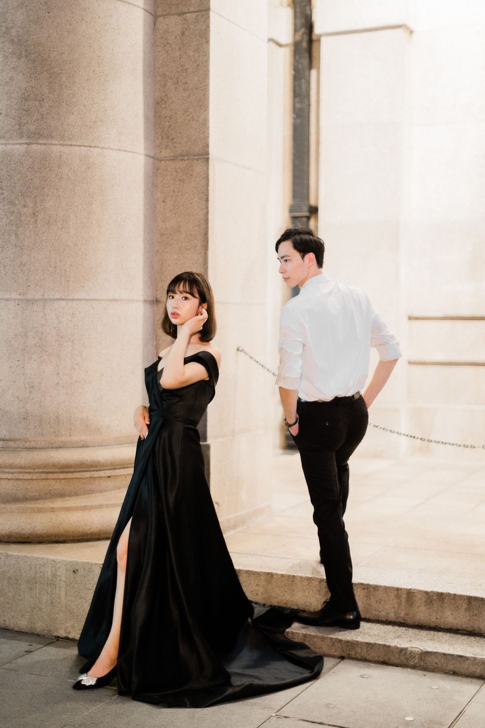 Elegant woman in black off-shoulder gown poses near stone columns with man in white shirt.