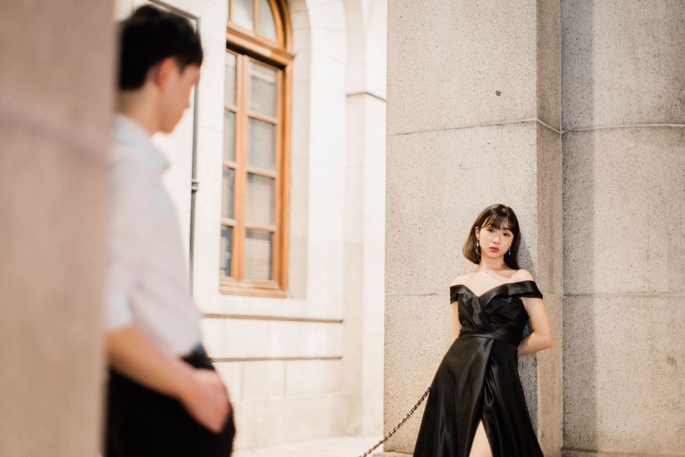 Asian couple in formal attire posing near stone building columns for pre-wedding photoshoot.