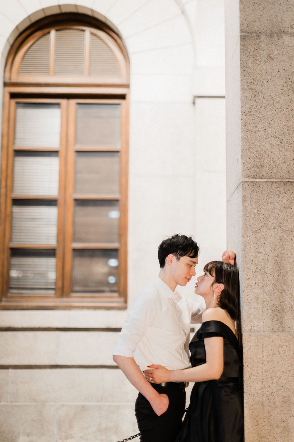 Romantic couple sharing intimate moment leaning against stone wall near arched window at dusk.