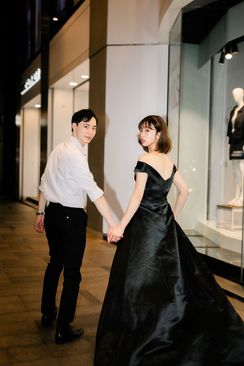 Couple holding hands at night outside luxury boutique, woman wearing elegant black ball gown.