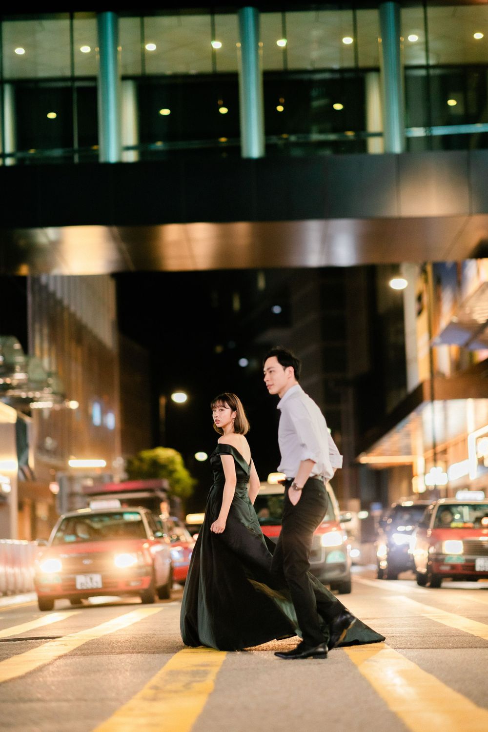 Couple in formal attire crossing a busy city street at night, woman in flowing black gown leads the way.