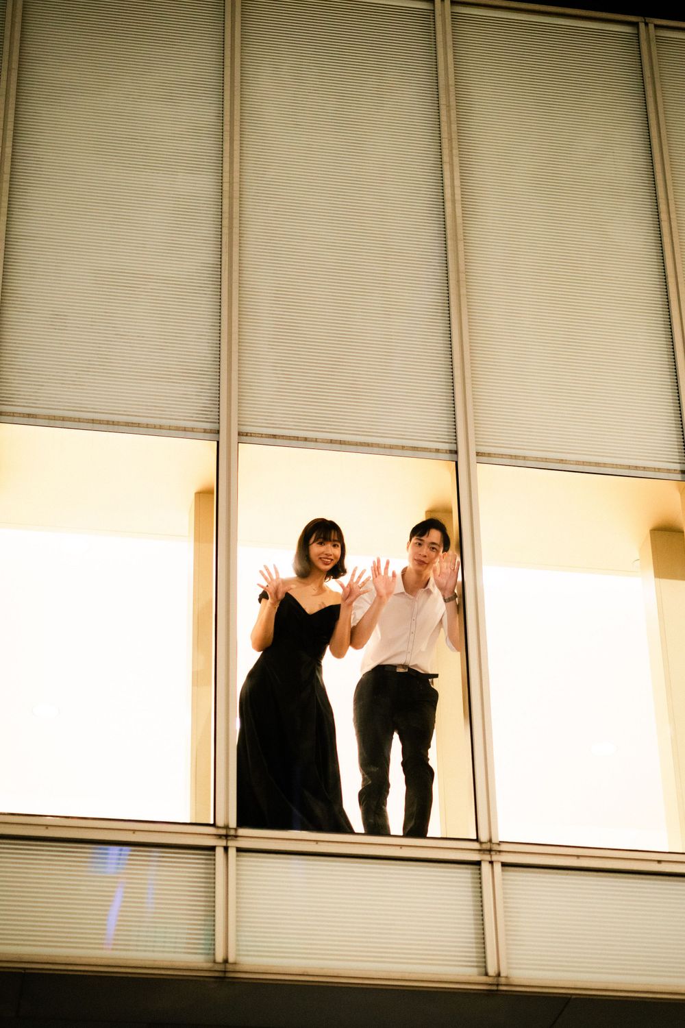 Playful couple pressing hands against a large illuminated window, smiling down from above at night.