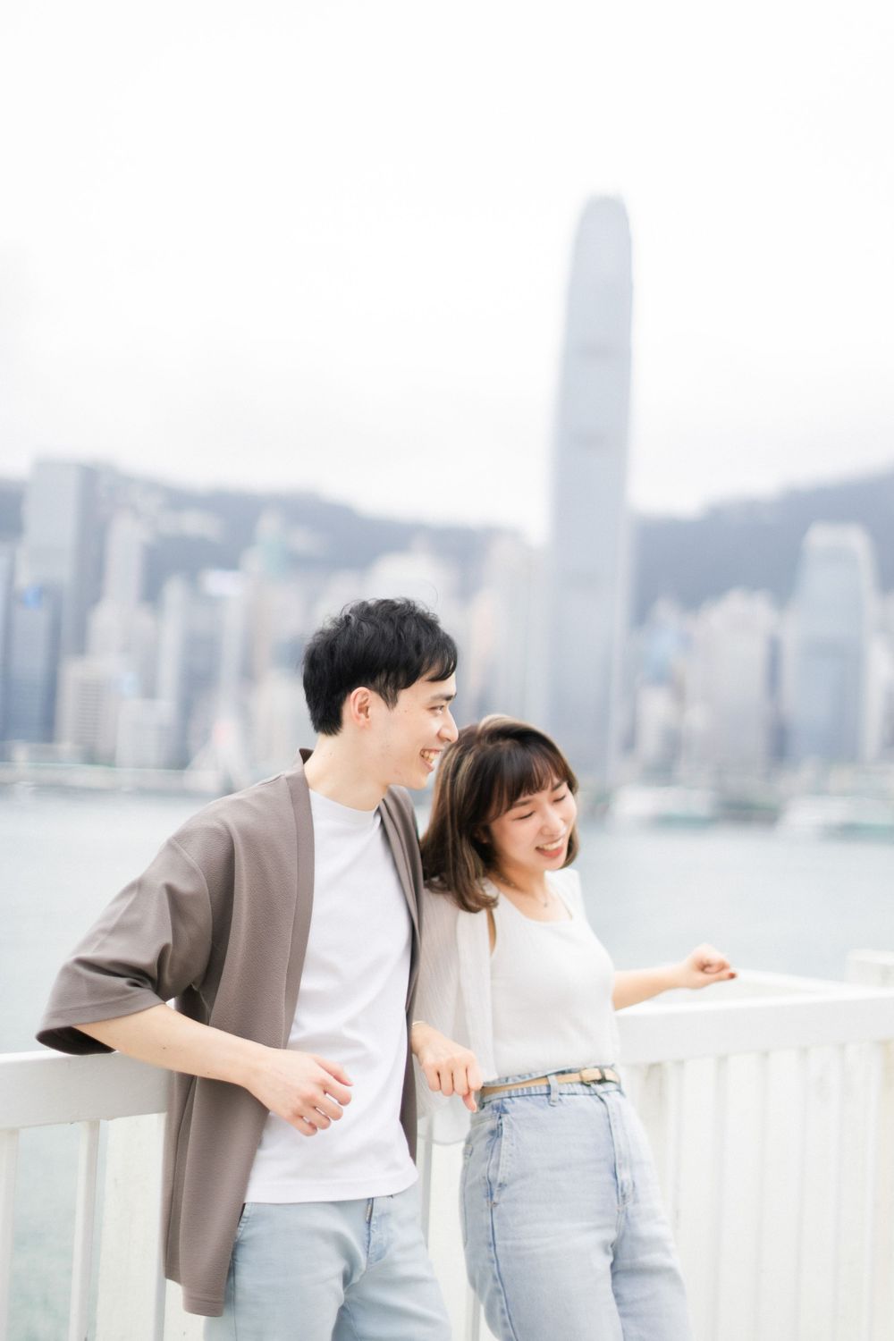 Happy couple leaning on white railing with Hong Kong Victoria Harbour skyline in the background.