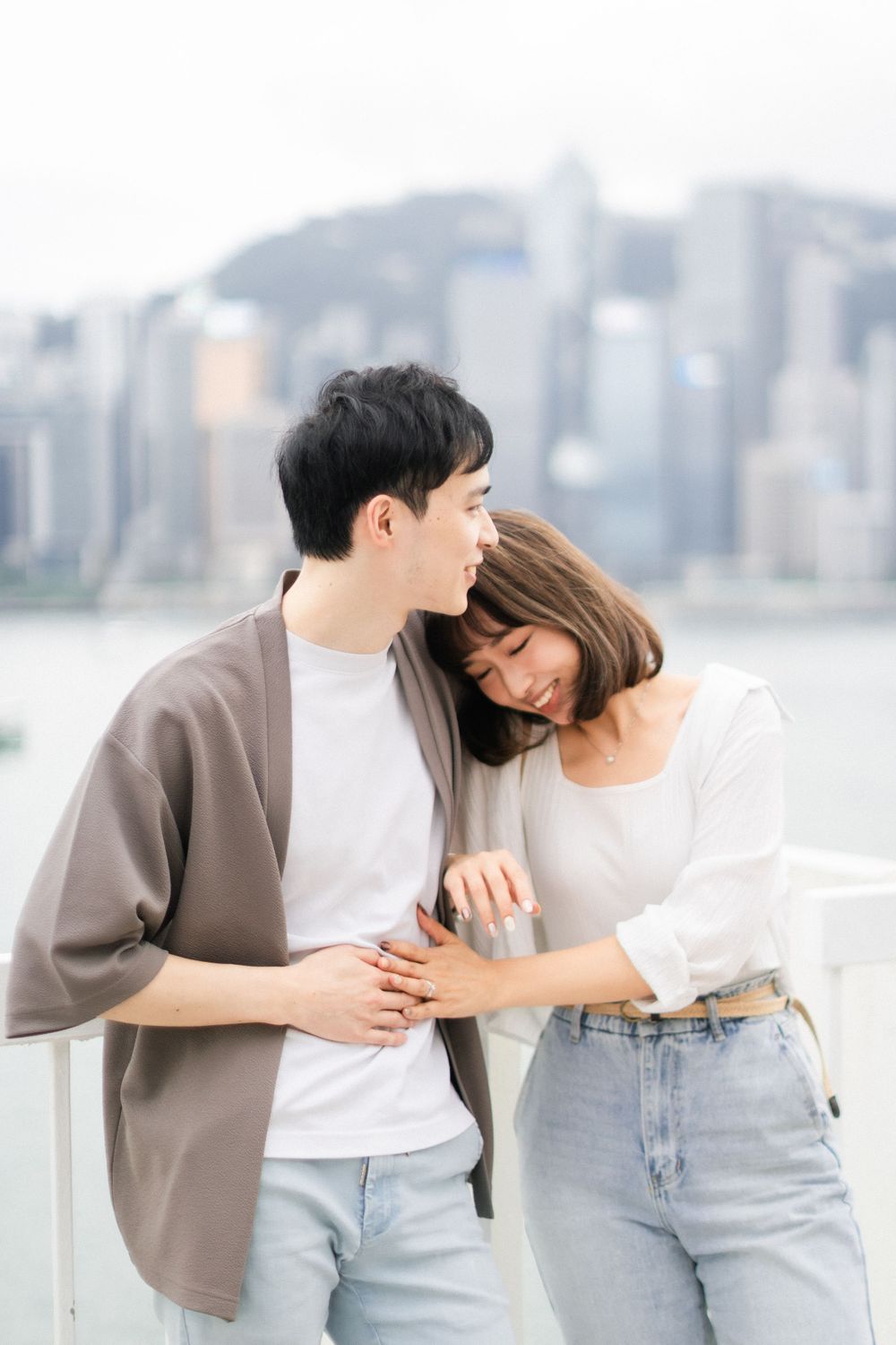 Couple sharing a tender moment by the waterfront railing with Hong Kong city skyline behind them.