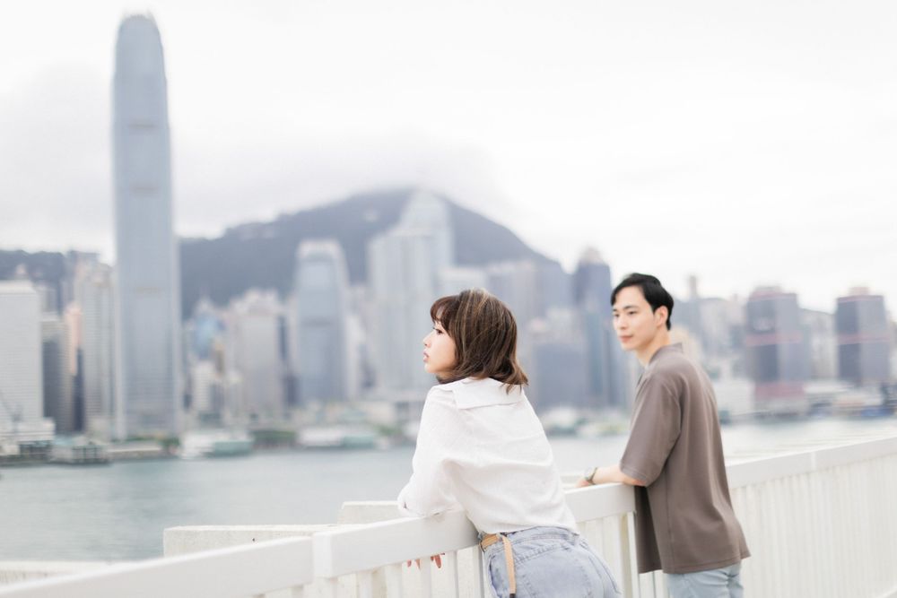 Young woman in white shirt leans on railing as man stands behind her with Hong Kong skyline backdrop.