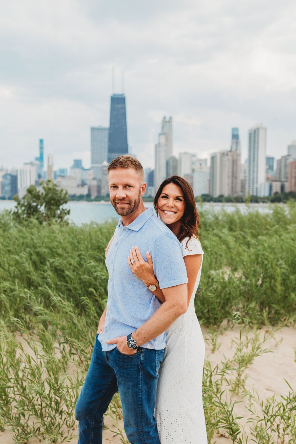 Couple shares joyful embrace on sandy beach with urban skyline backdrop.