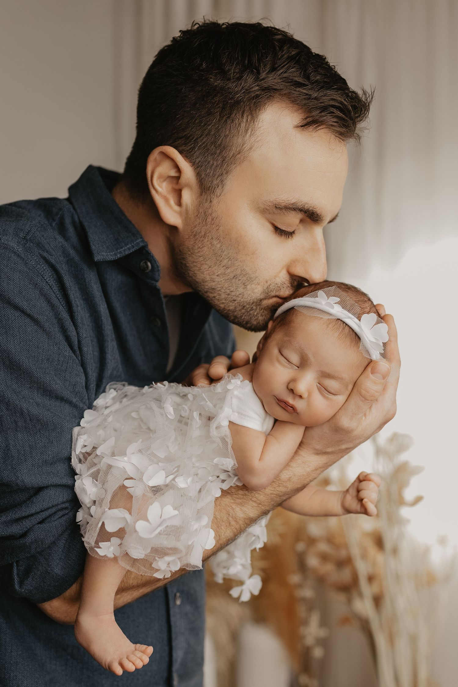 A father tenderly kisses his sleeping newborn baby girl in a white floral dress and headband.