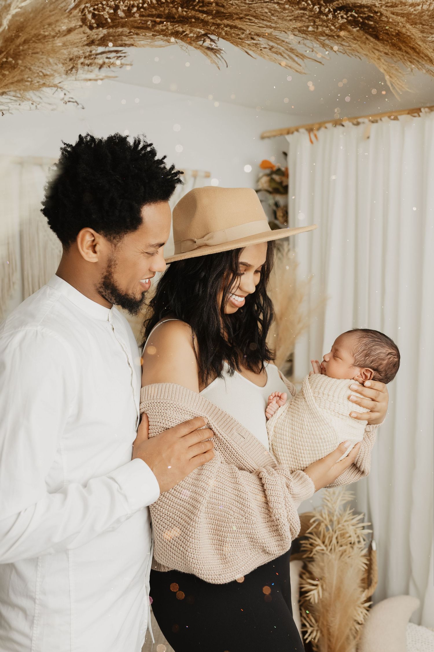 Stylish couple in neutral tones holding their newborn in boho-decorated room with pampas grass.