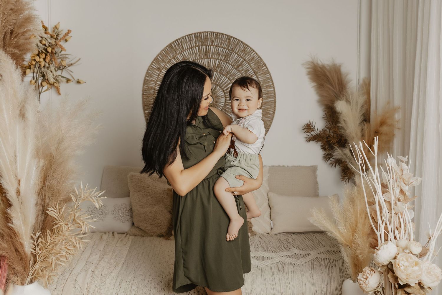 A mother in an olive dress holds her baby in a boho-styled room with pampas grass and warm neutral tones.