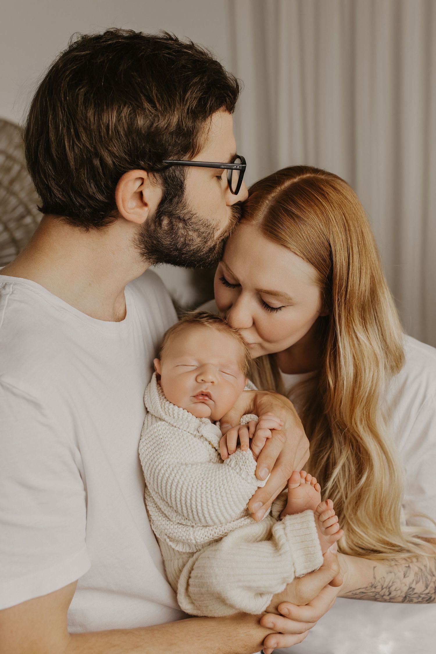 Parents kissing their sleeping newborn baby wrapped in a knit blanket, a tender family portrait in soft neutral tones.