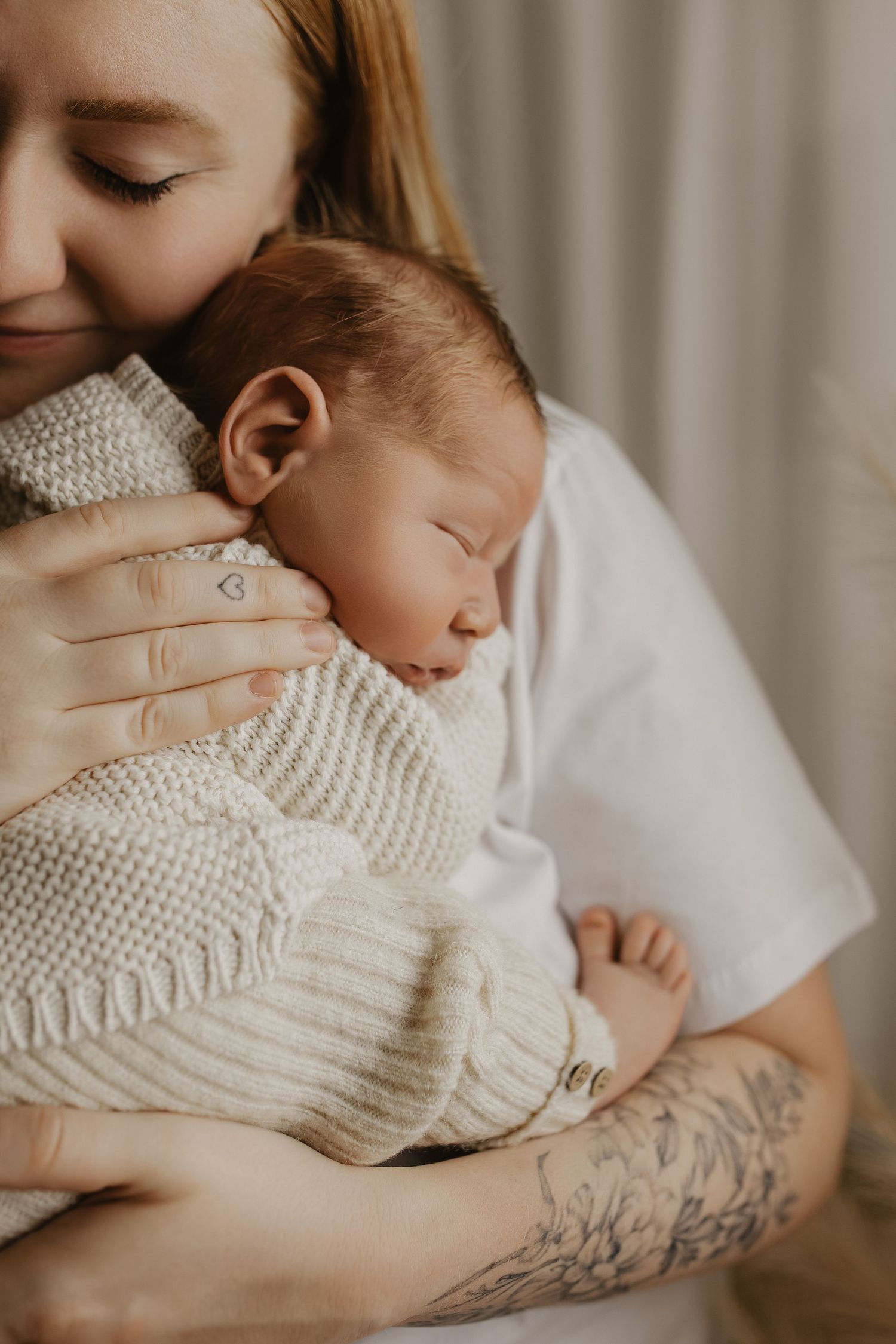 A tattooed mother tenderly holds her sleeping newborn baby wrapped in a cozy cream knit blanket.