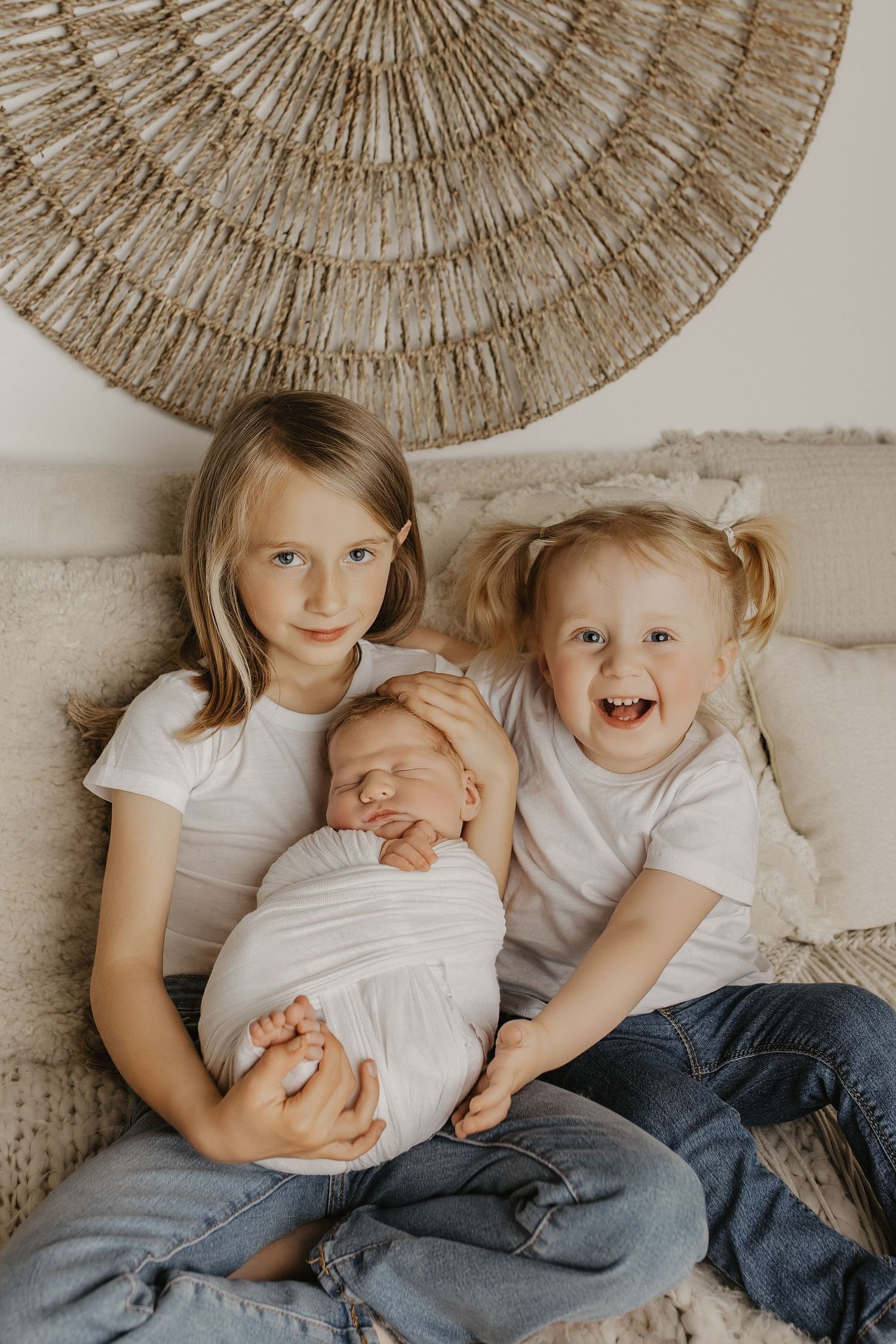 Two older sisters in white tees and jeans lovingly hold their swaddled newborn sibling on a cozy neutral-toned sofa.