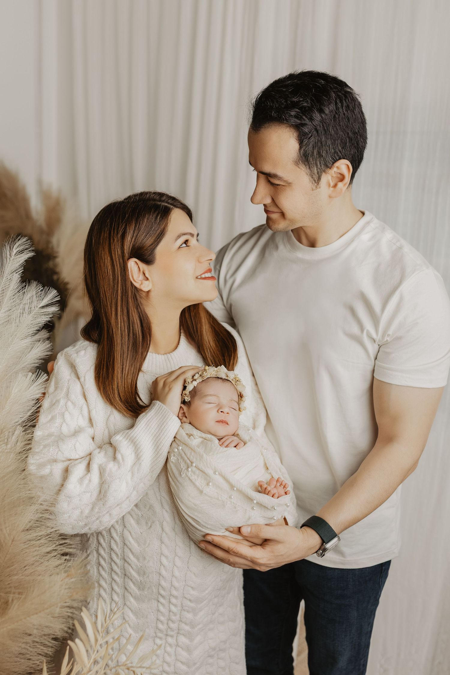 A couple smiles at each other while tenderly holding their sleeping newborn baby wrapped in a white blanket.