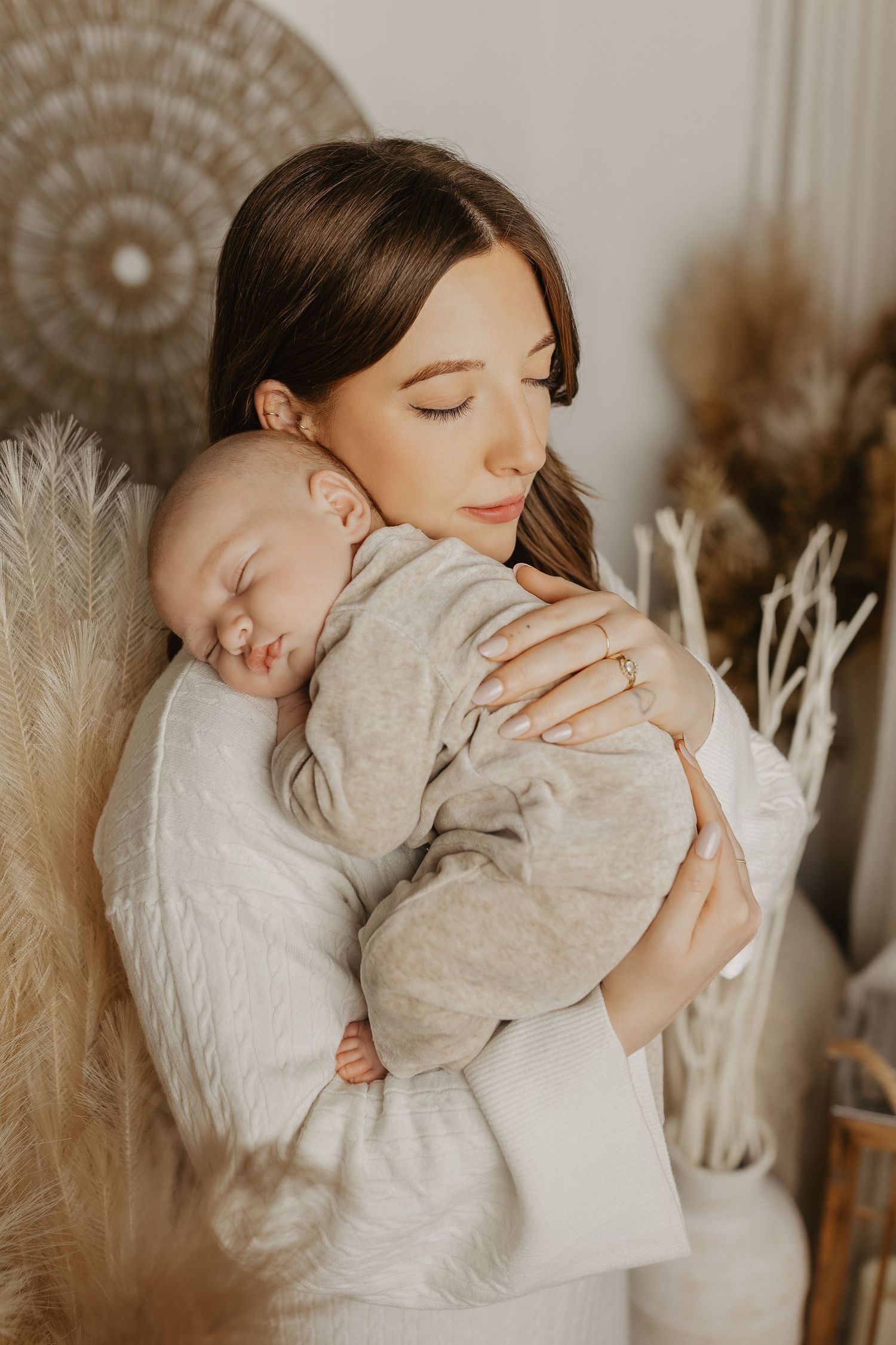 Parent in white sweater cuddling sleeping baby against neutral backdrop with pampas grass decor.
