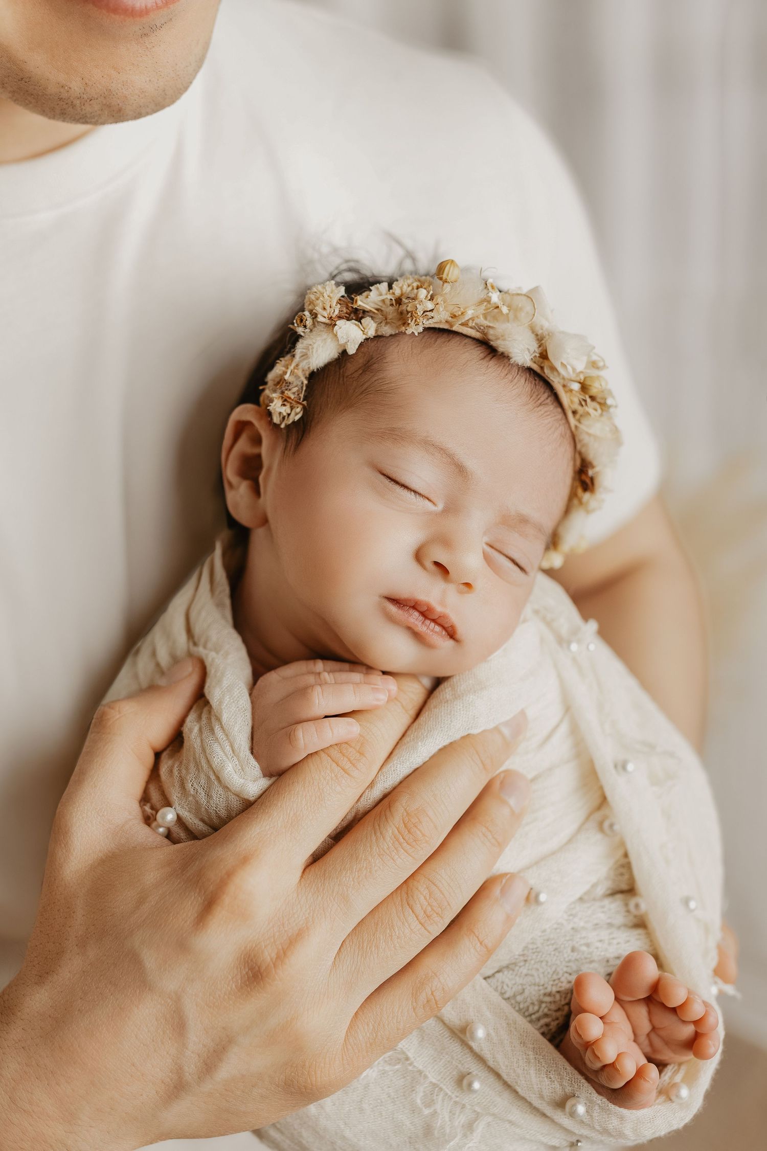 Sleeping newborn baby girl wearing a floral crown, swaddled in cream wrap, cradled in father's hands.