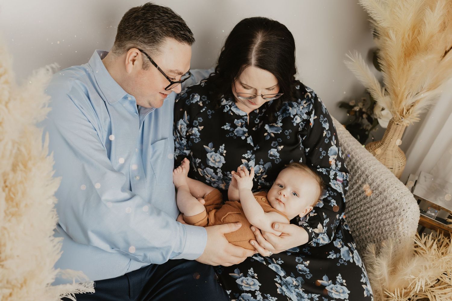 Family sitting on wicker chair sharing tender moment with baby surrounded by pampas grass decor.