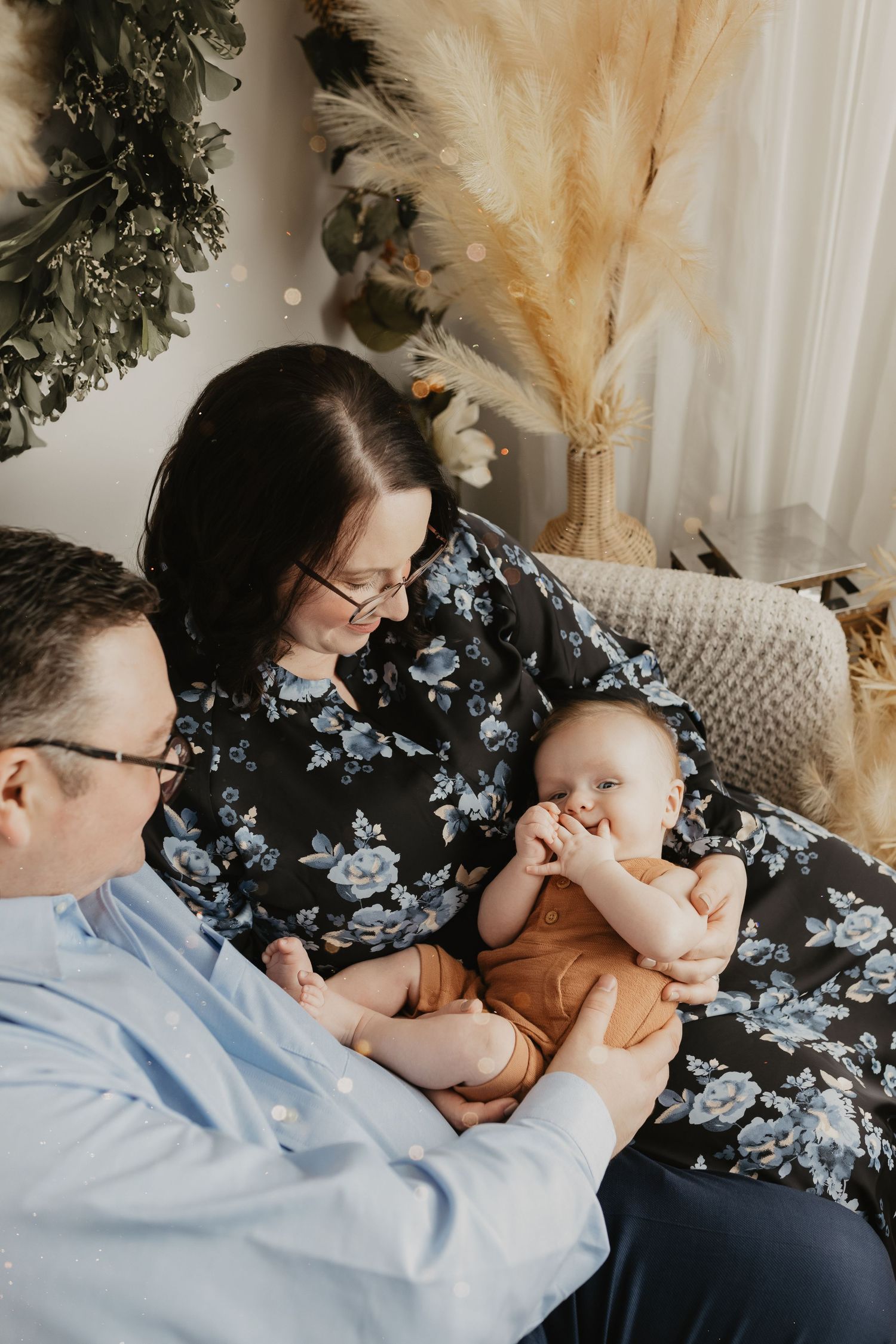 Candid family moment on wicker chair with baby surrounded by dried floral arrangements.