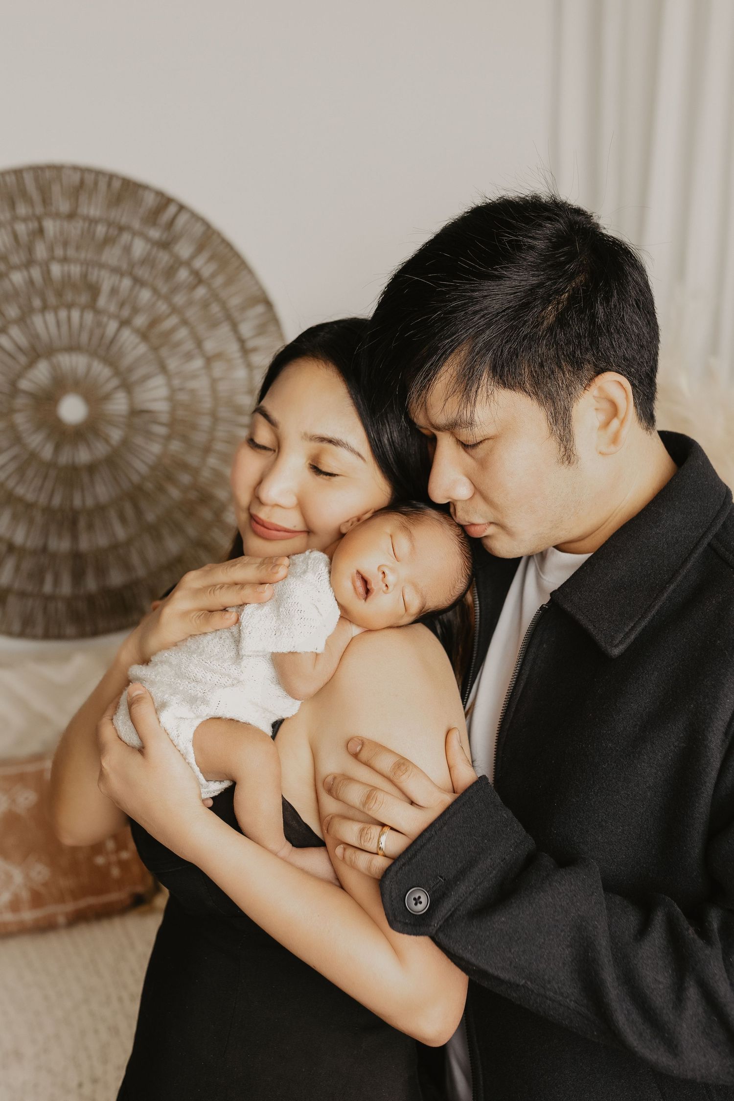 A tender family moment showing a parent in black cradling and kissing a sleeping baby in white against a neutral backdrop.