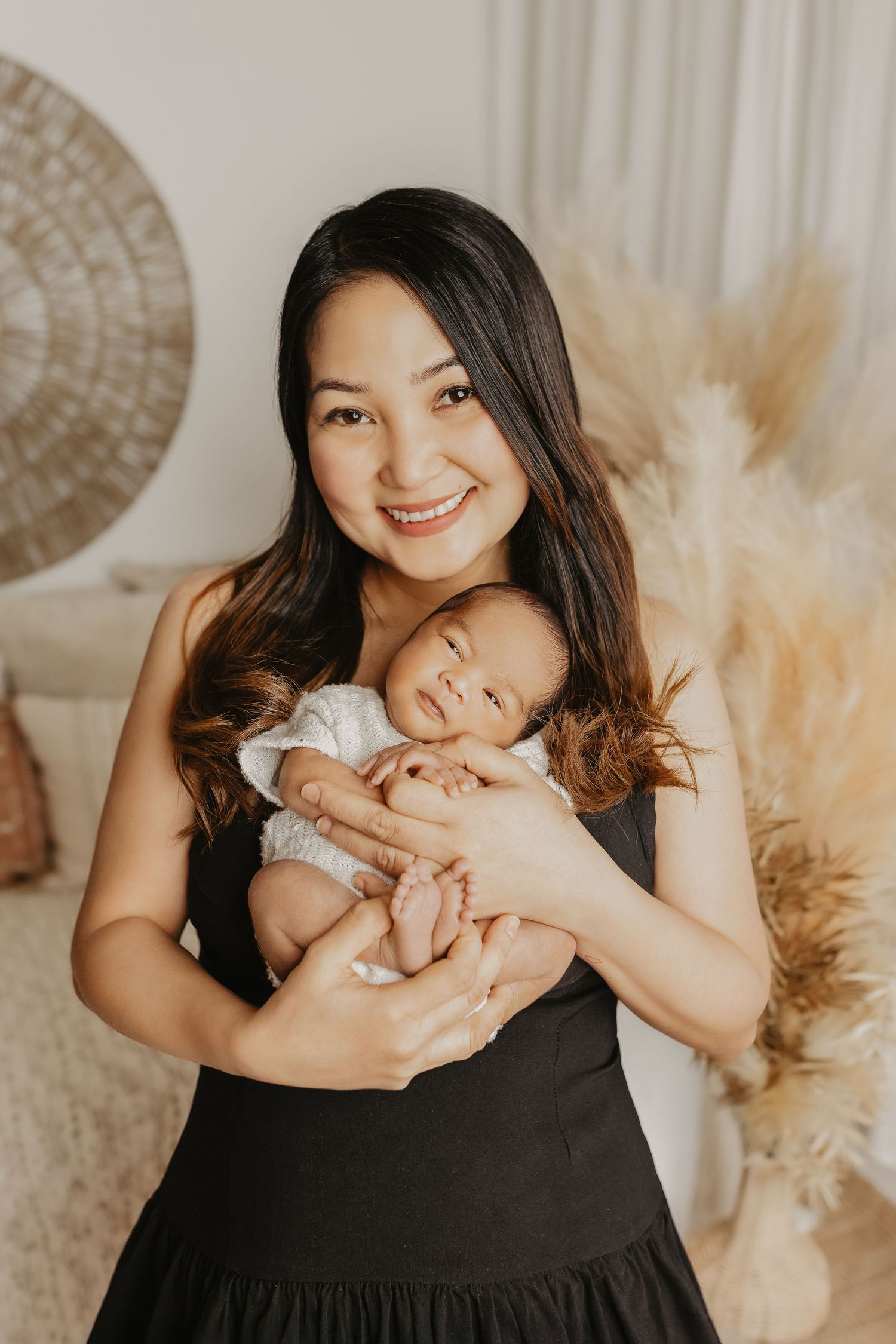 A mother in a black dress holds her sleeping newborn baby in a cozy room decorated with pampas grass and woven decor.