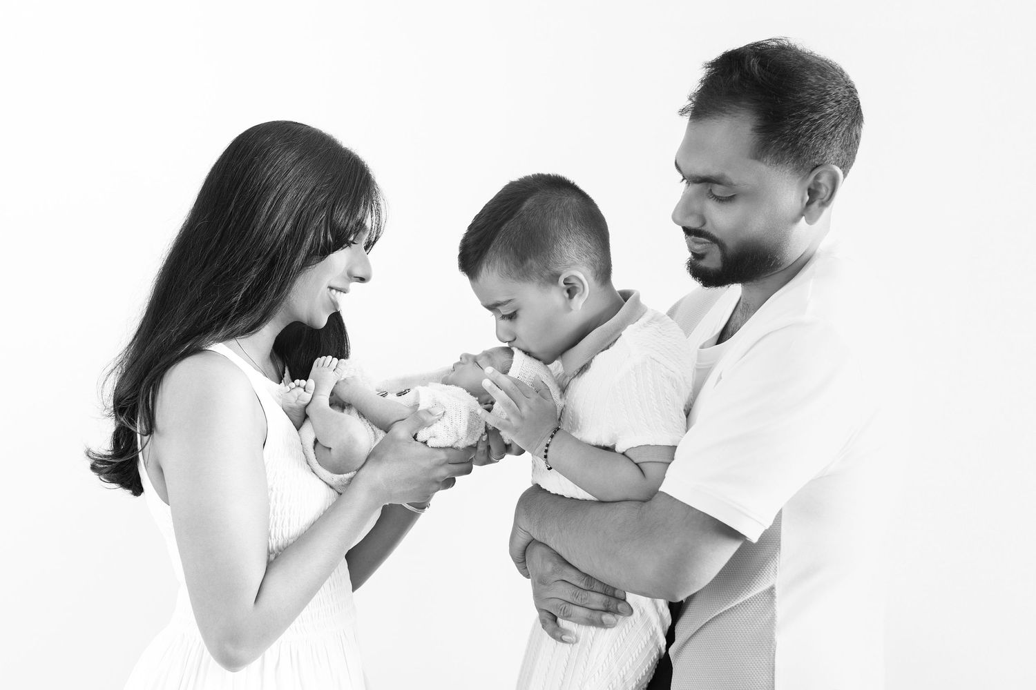 Black and white family portrait of parents with toddler son and newborn baby in a studio setting.
