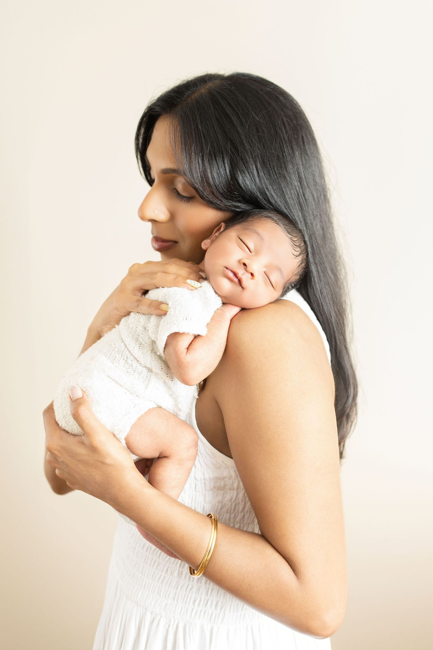 A mother in a white dress tenderly holds her sleeping newborn baby against her shoulder, wearing a gold bracelet.