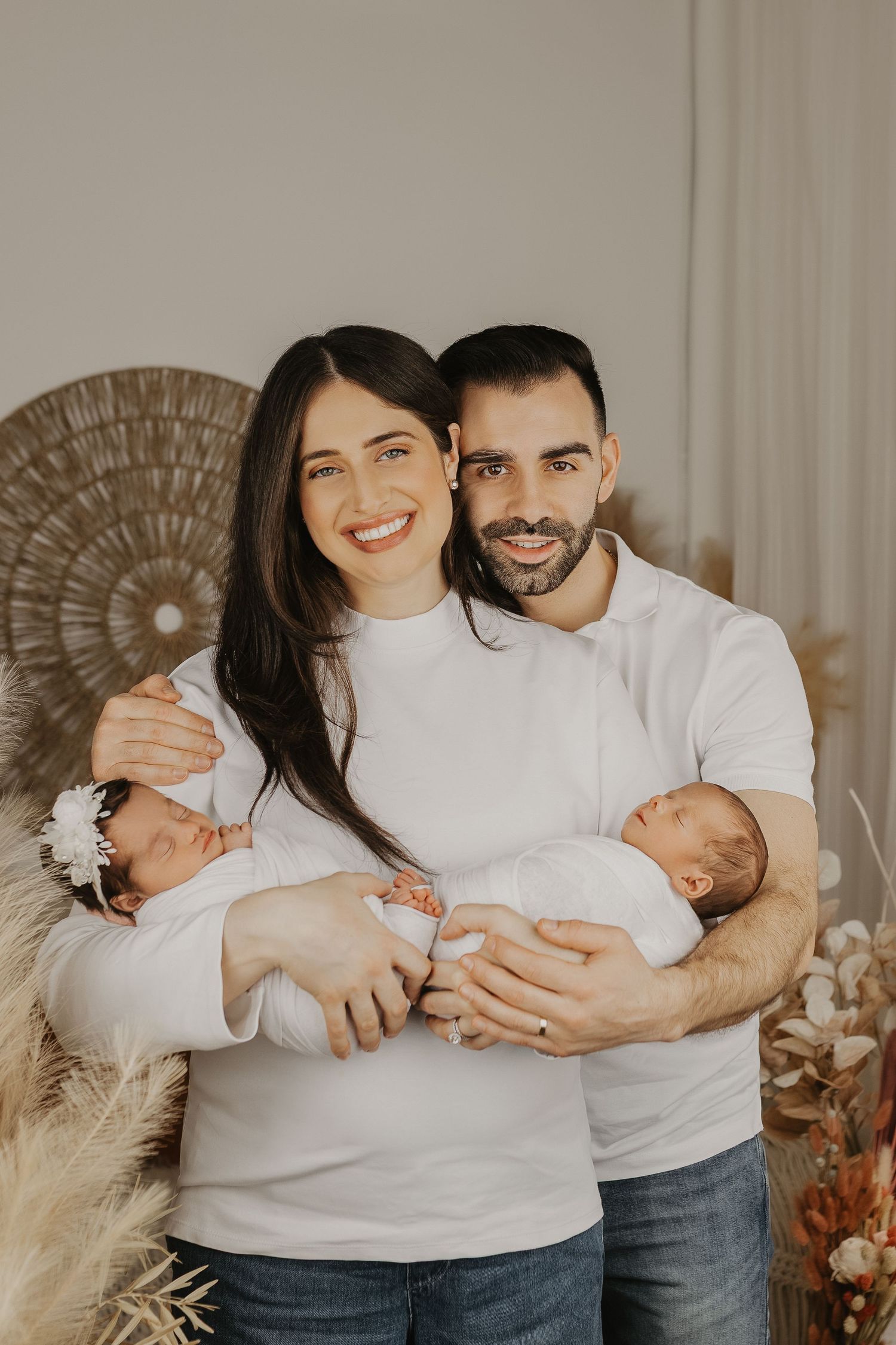 Smiling couple in white tops holding newborn twins in a cozy studio with boho decor and dried floral arrangements.