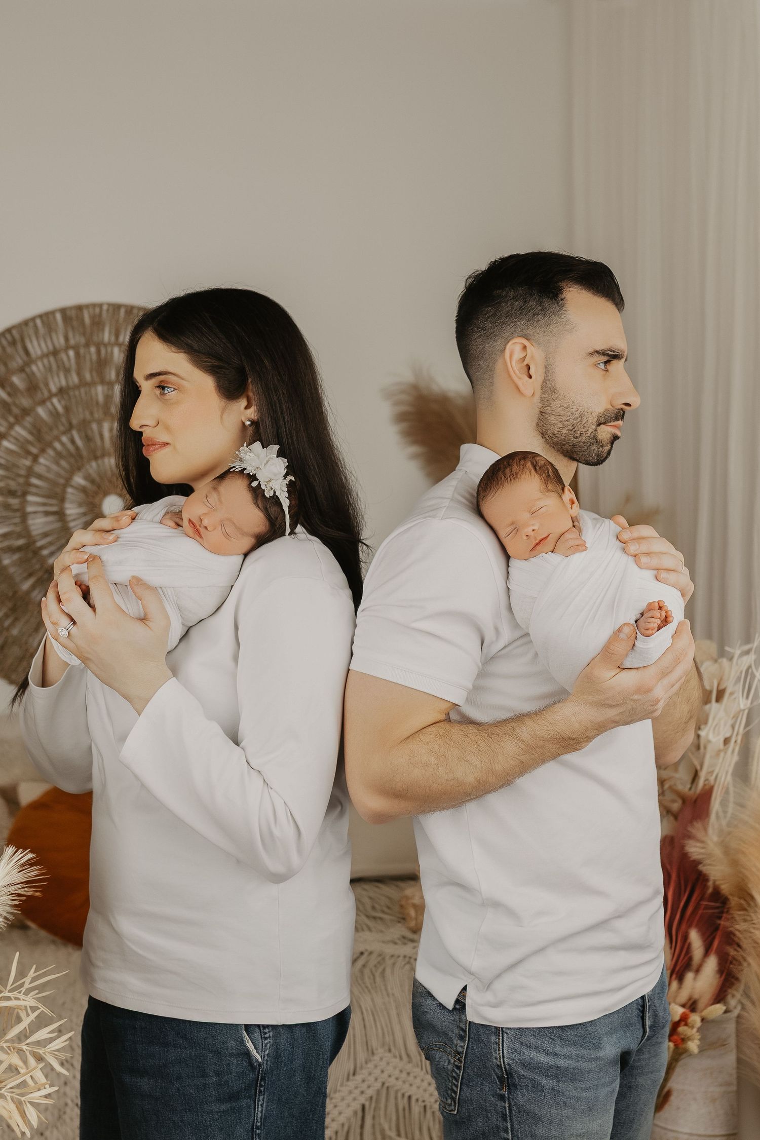 A couple in white shirts stand back-to-back, each cradling a sleeping newborn baby in a soft, bohemian-styled room.