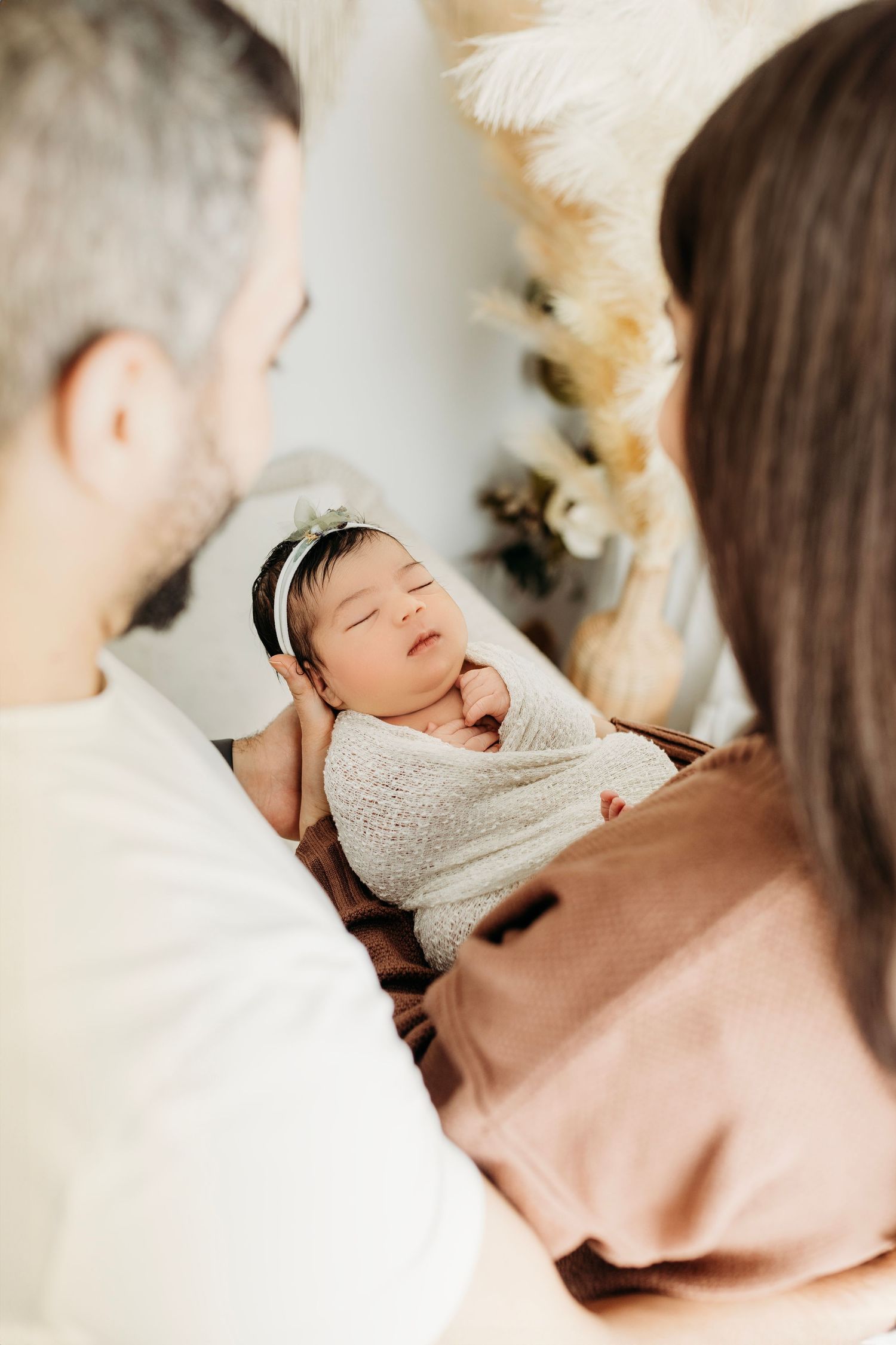 Parents gazing lovingly at their sleeping newborn baby wrapped in white swaddle.