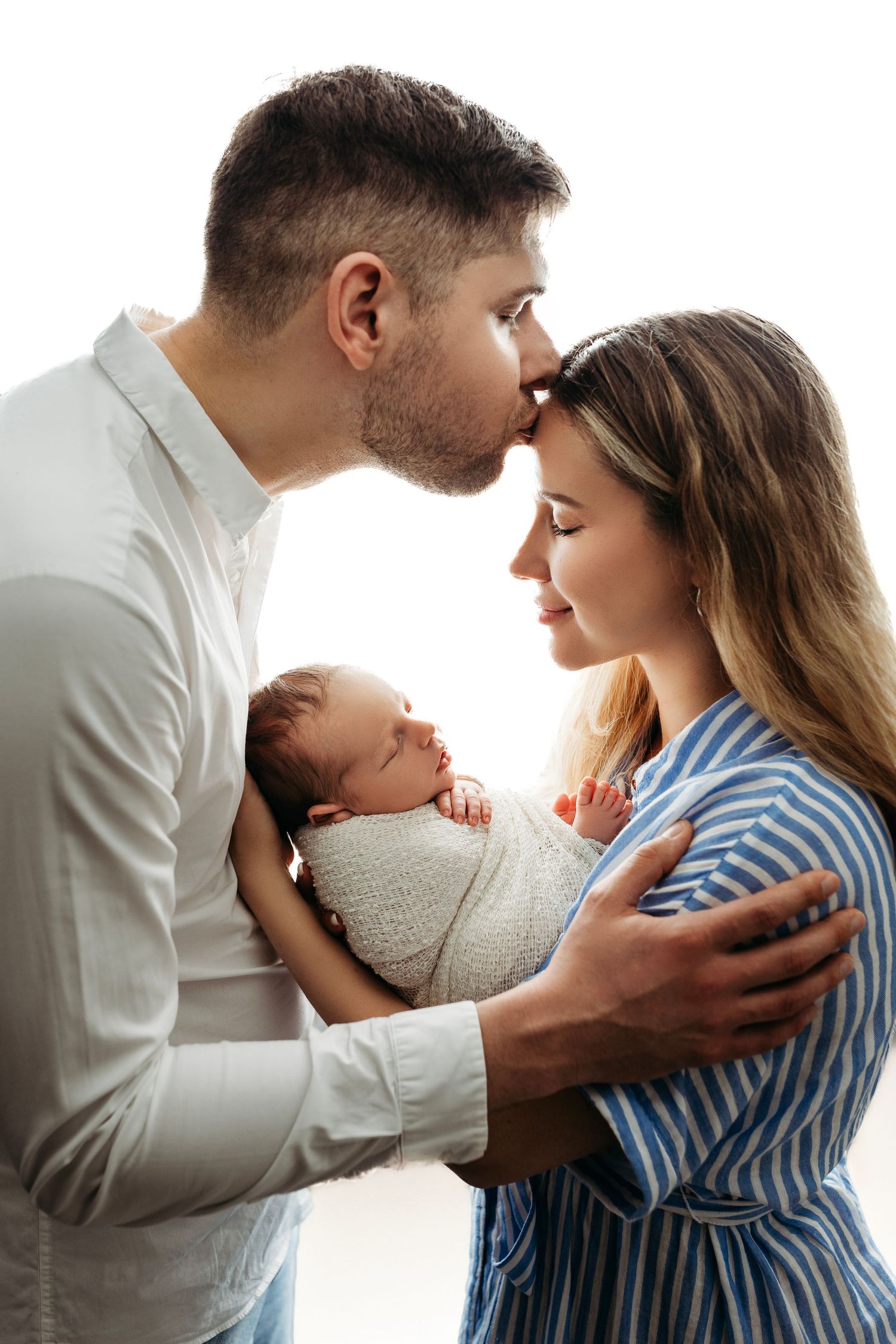 A tender family moment as parents embrace while holding their sleeping newborn baby wrapped in white.