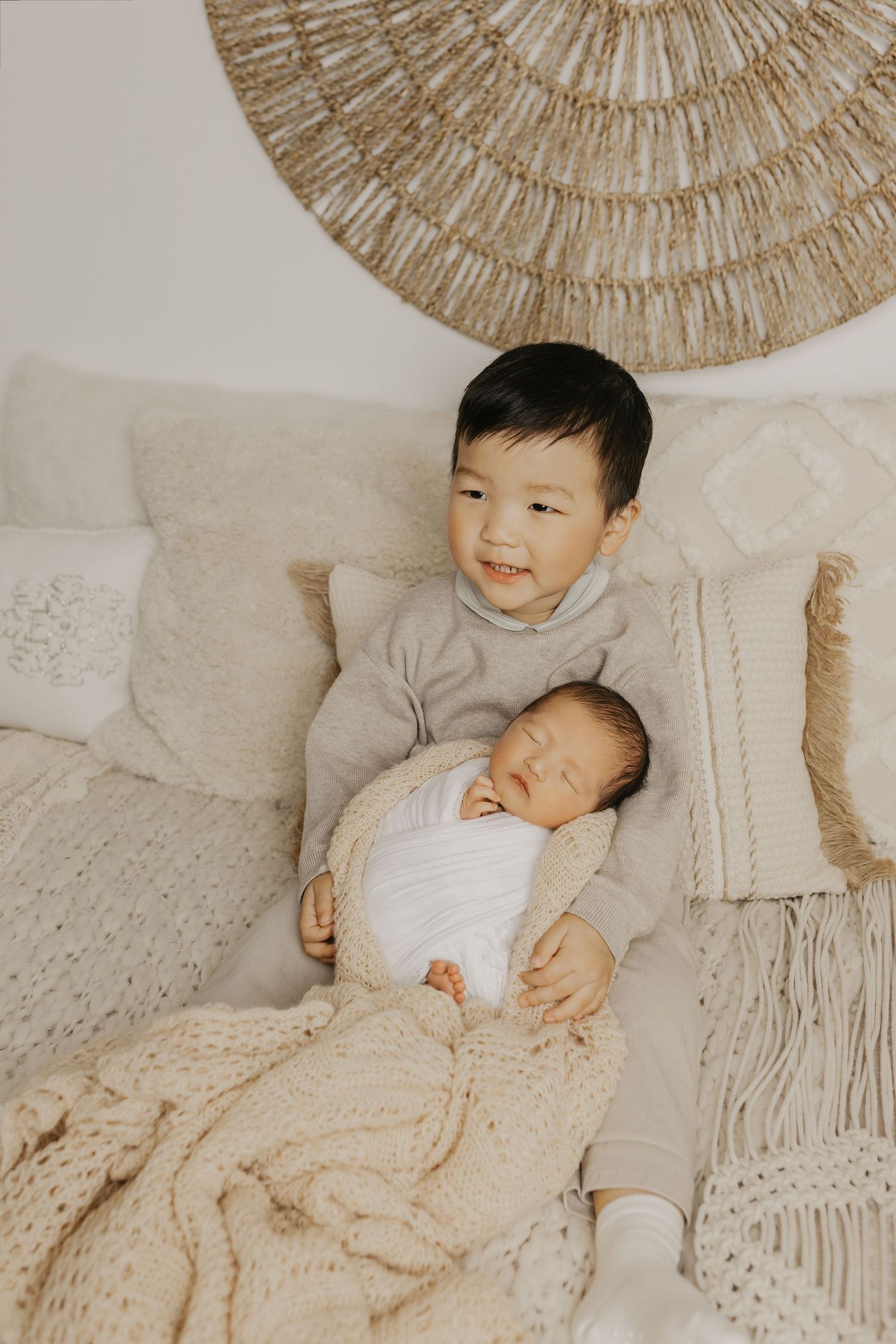 Young boy in gray sweater gently holds sleeping swaddled newborn baby on cozy cream knit blanket.