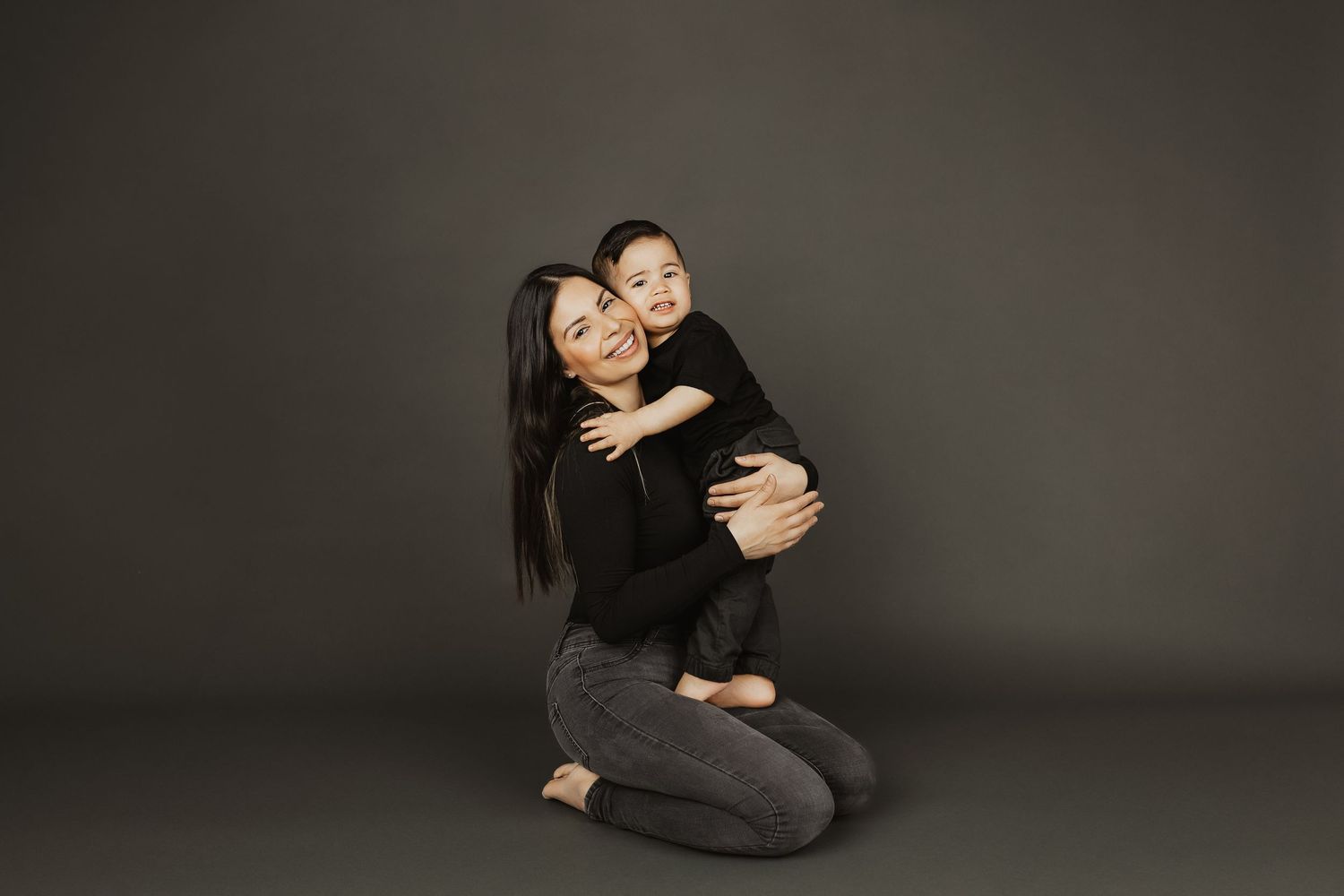 A mother kneeling on the floor hugging her toddler, both dressed in black, smiling against a gray background.
