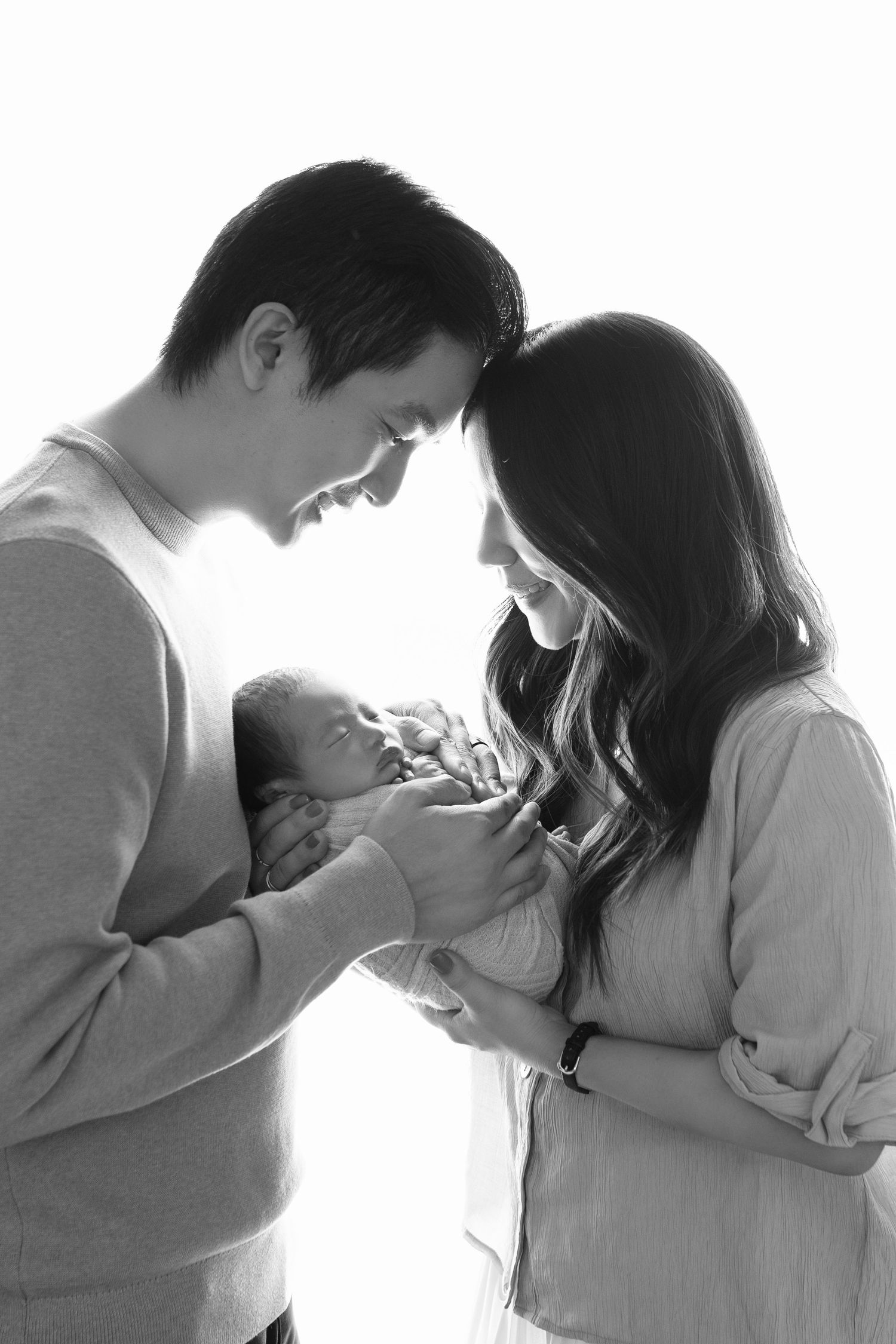 Black and white photo of smiling parents lovingly gazing at their newborn baby held between them.