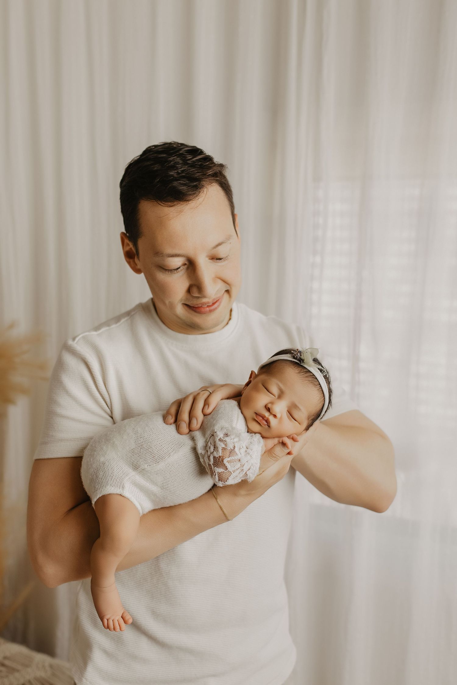 A smiling father in a white t-shirt tenderly holds his sleeping newborn baby girl near a bright window.