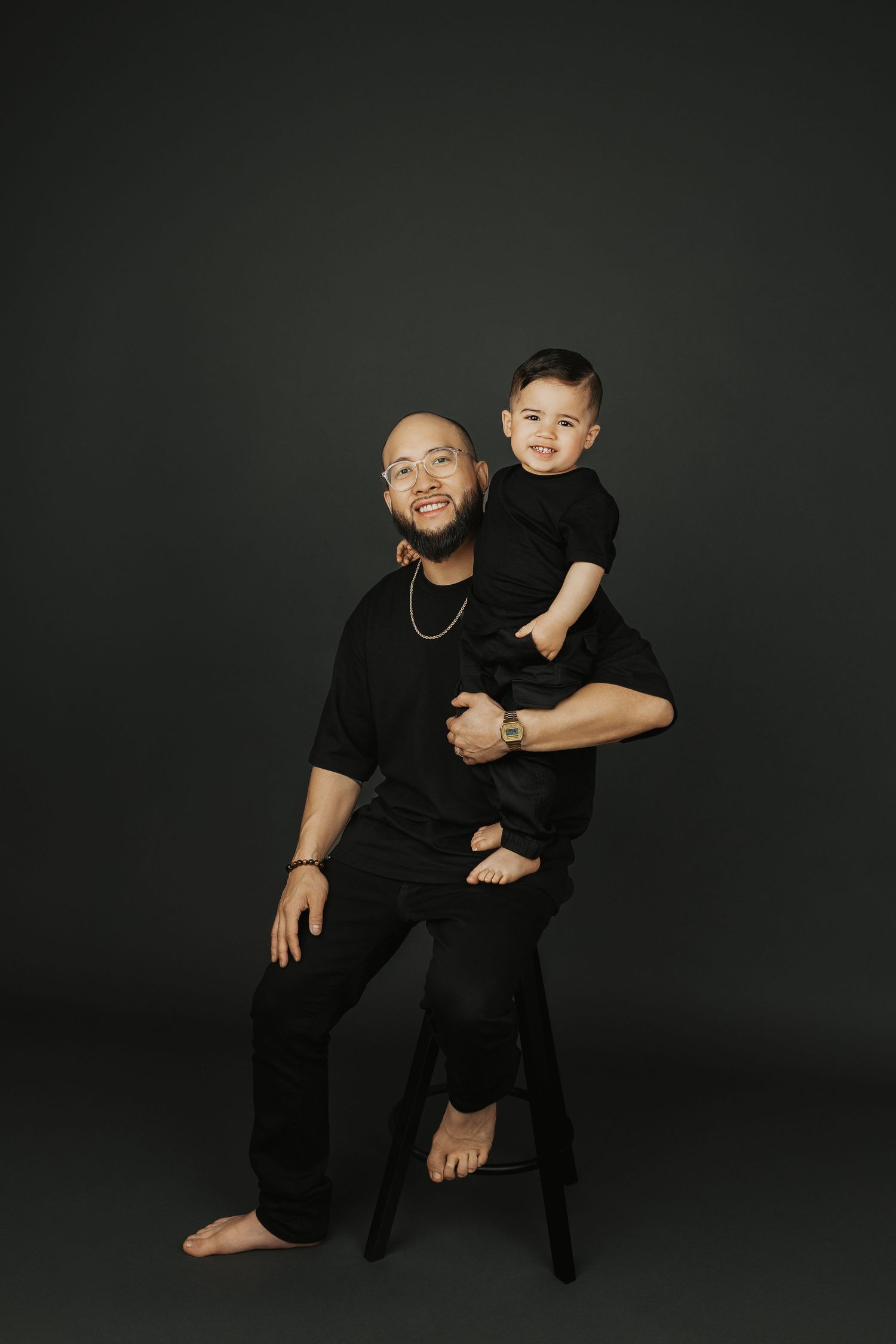 A bald man in black outfit sits on a stool holding a young boy dressed in matching black clothes against dark background.