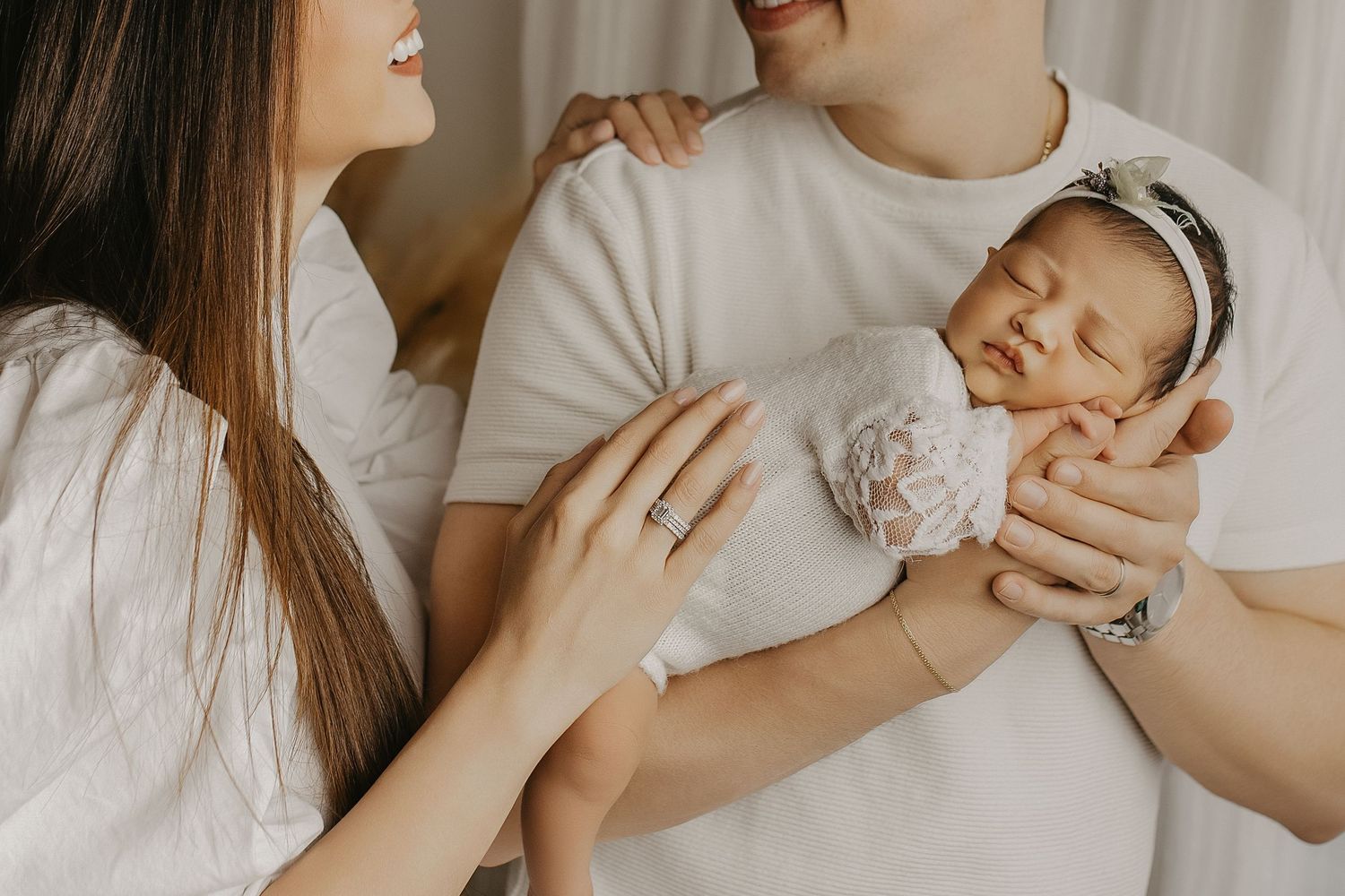 New parents in white shirts lovingly hold their sleeping newborn baby wrapped in a delicate white lace outfit.