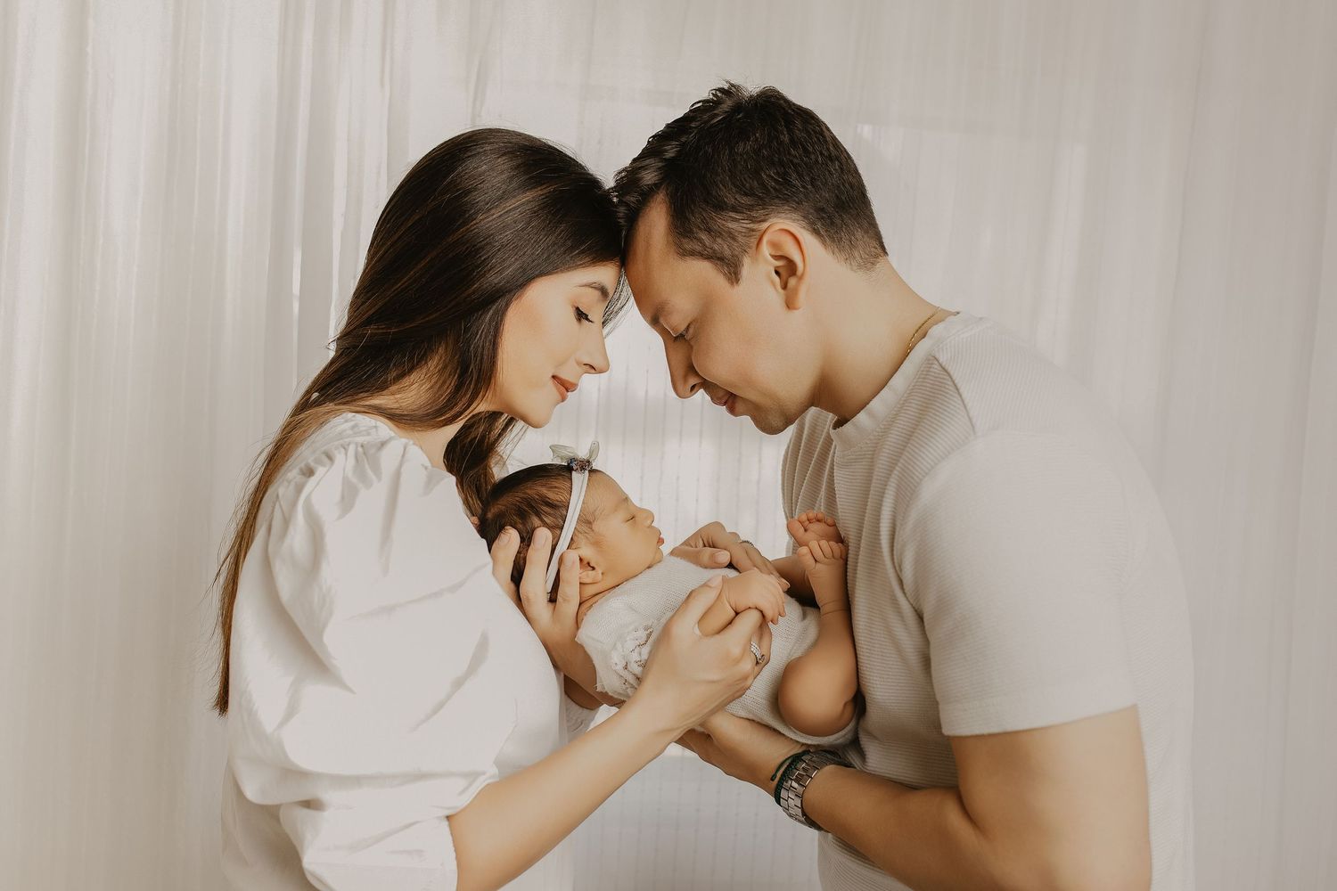 A couple tenderly holds their newborn baby together, touching foreheads, against a soft white curtain backdrop.