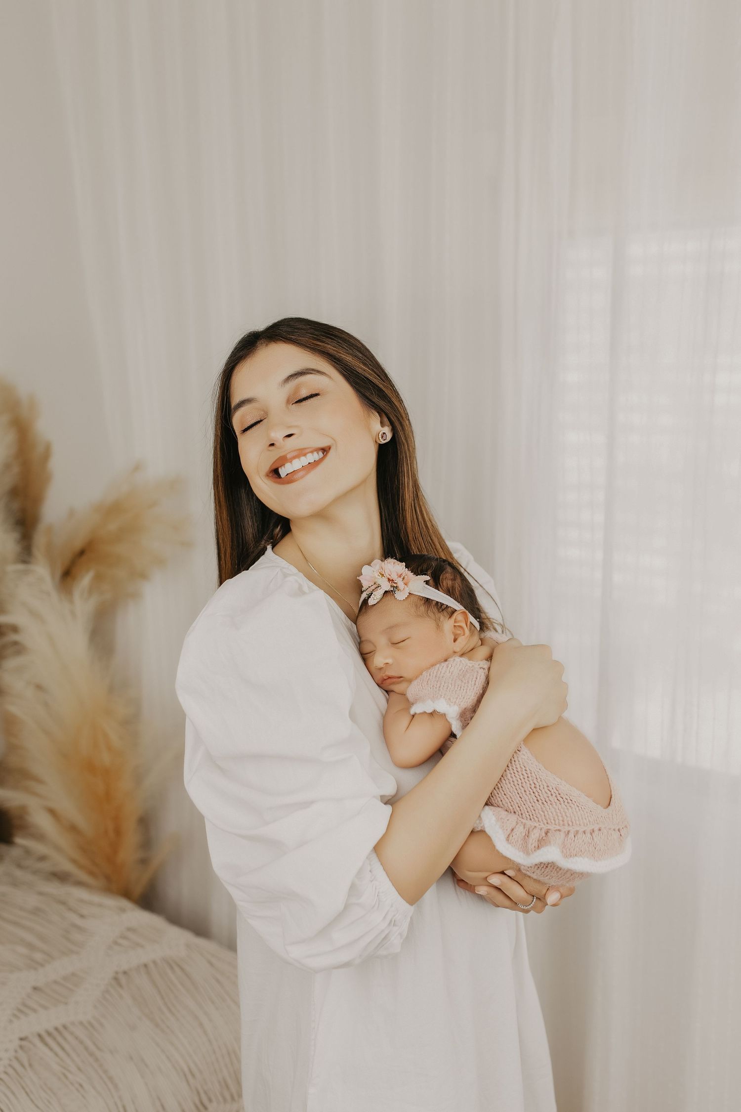 A smiling young mother in a white dress tenderly holds her sleeping newborn baby girl wearing a pink floral headband.